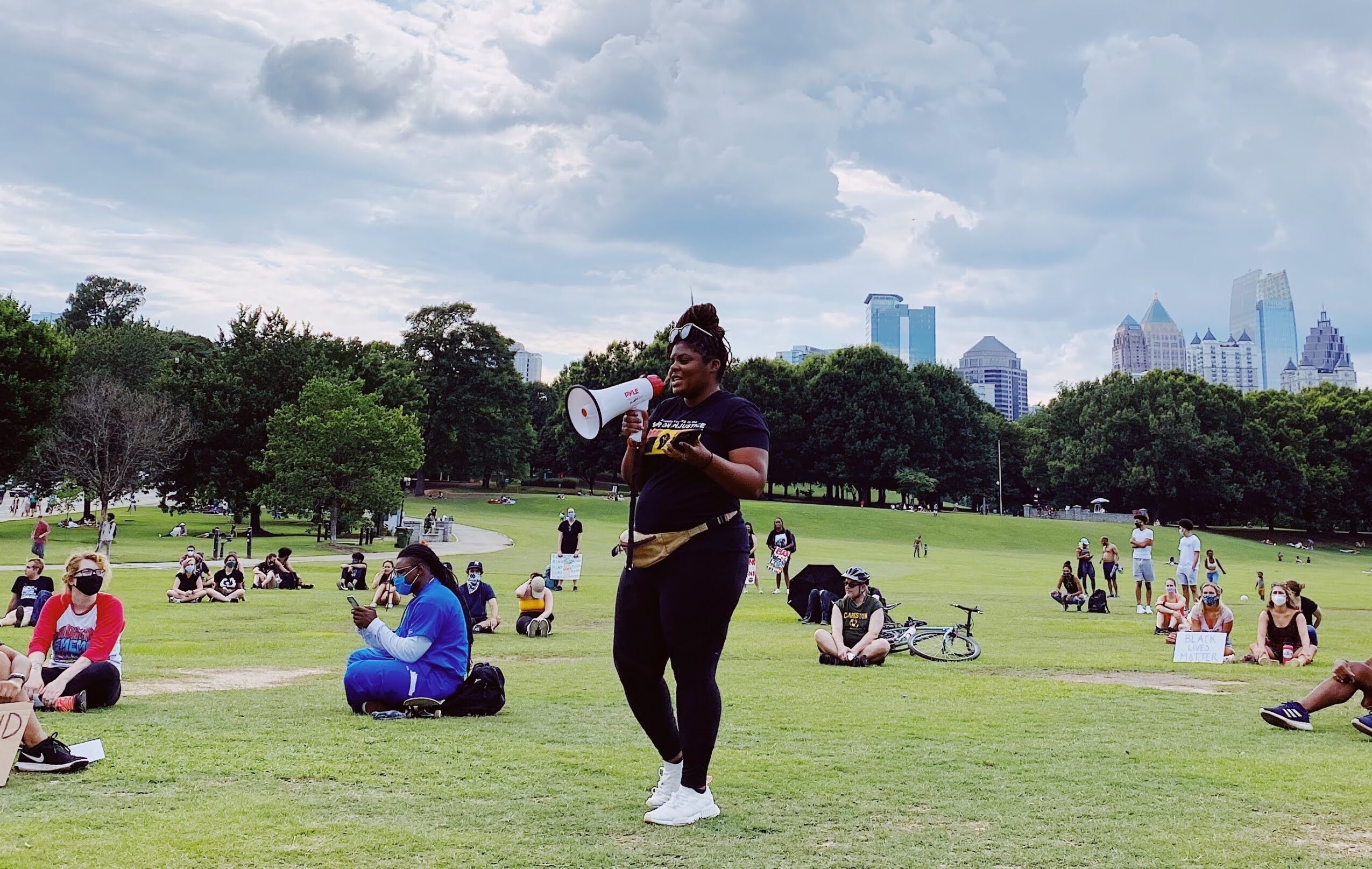 Person standing on grass holding a megaphone in a park with people sitting and walking, city skyline in background.