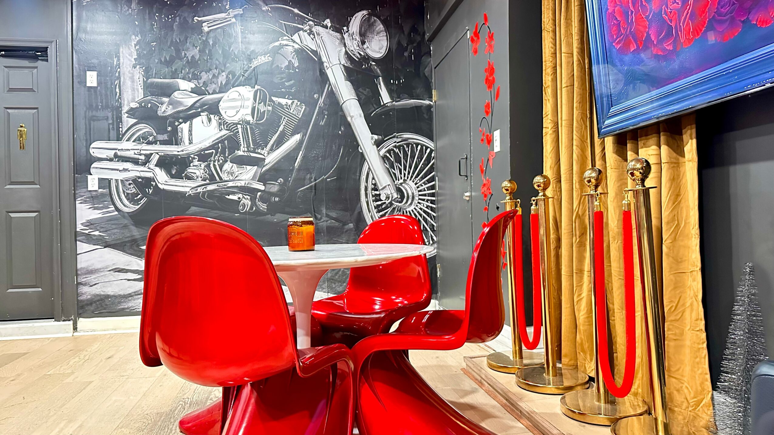 Red chairs around a round table in front of a black and white motorcycle mural, with gold stanchions and red ropes nearby.