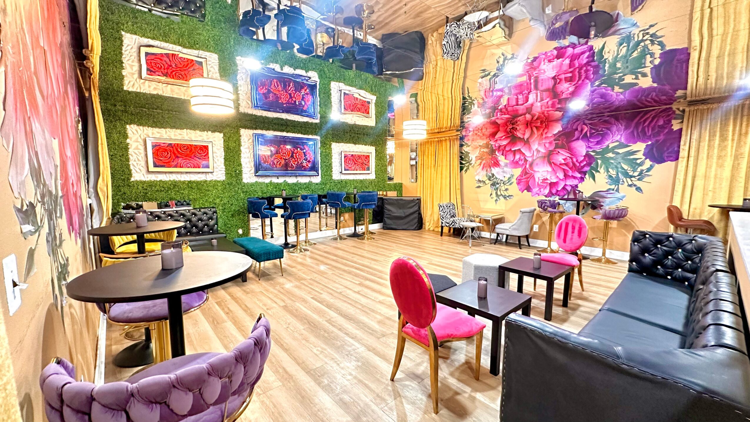 Room with colorful furniture, large floral painting, and multiple TV screens on the wall, with wooden flooring and yellow curtains.