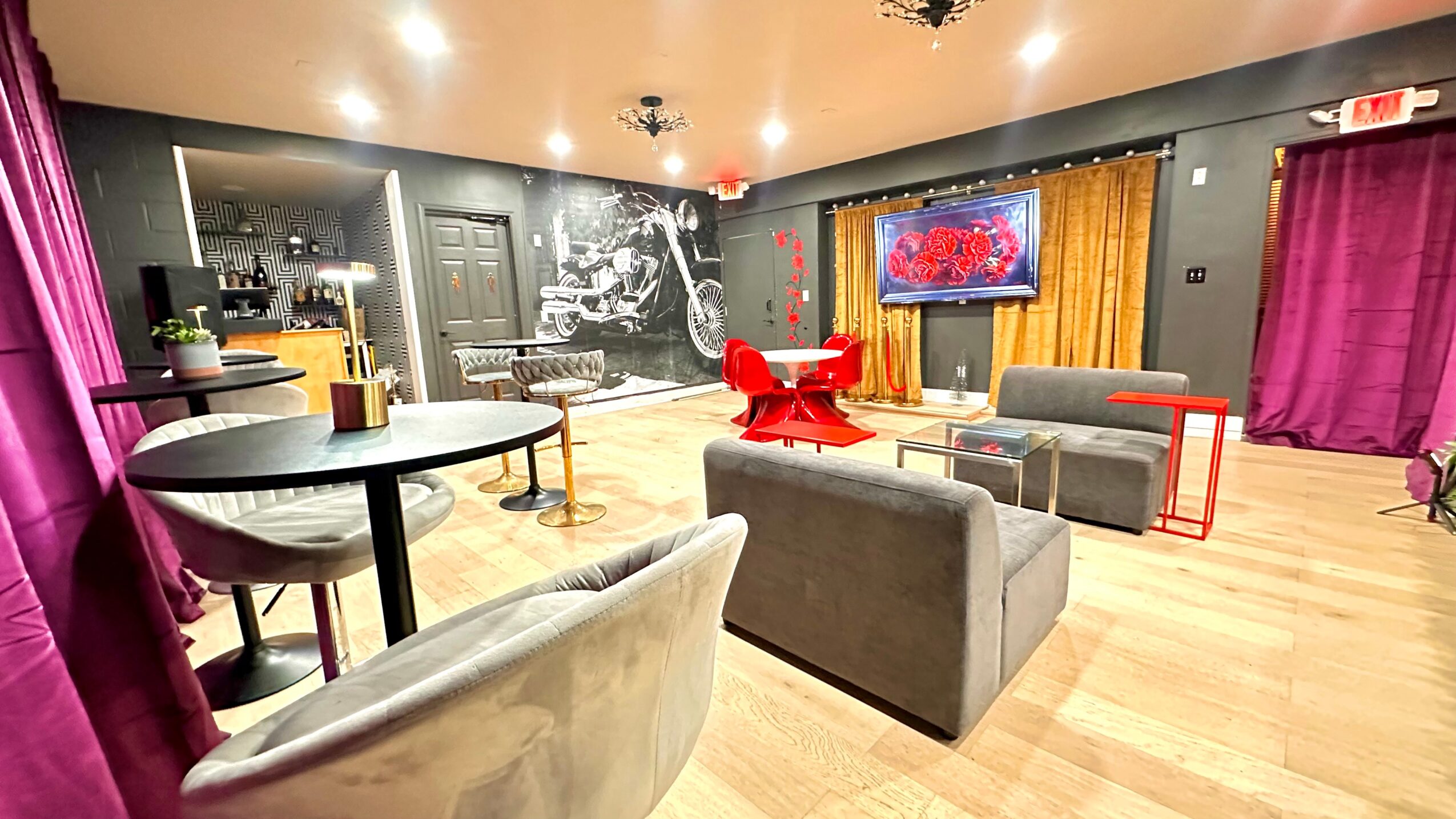 Living room with sofas, a TV, curtains, and a chalkboard wall, illuminated by ceiling lights, with wooden flooring.