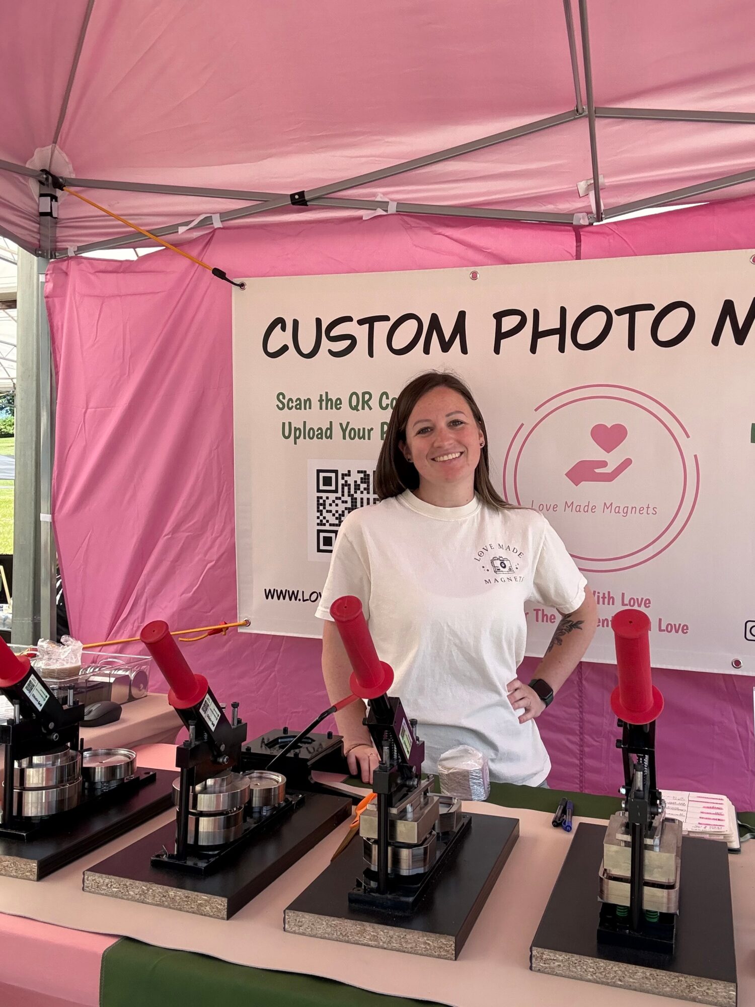 Smiling woman at a booth with custom photo mugs, pink tent, and a sign behind her, smiling and wearing a white shirt.