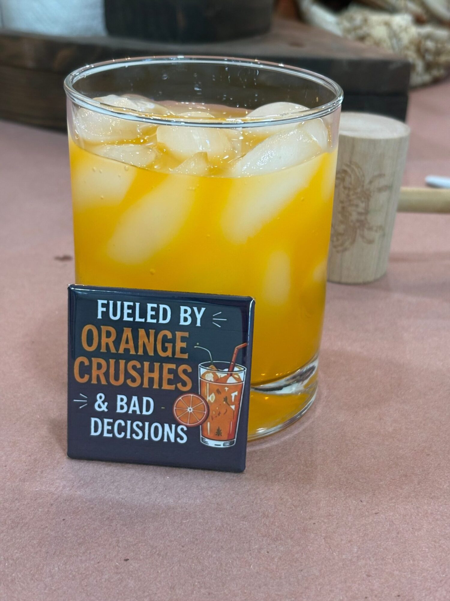 A glass of yellow and white layered drink with ice, placed on a pink surface, with a tag reading 'Fueled by Orange & Bad Crushed Decisons'.