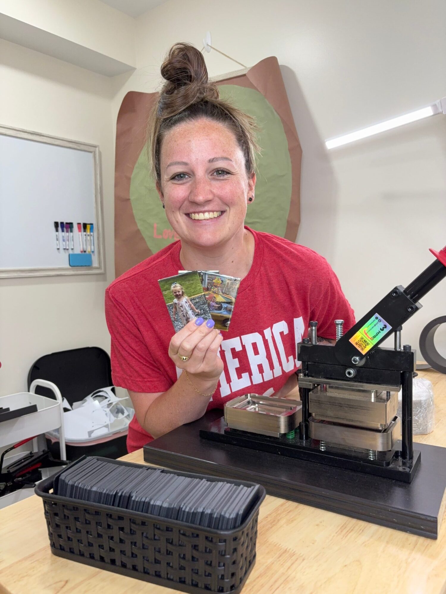 Woman smiling and holding a colorful object, sitting at a table with a 3D printer, in a room with a whiteboard.