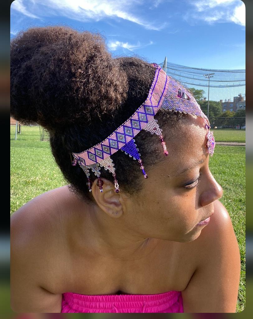 Woman with styled hair and beaded headpiece outdoors, looking to the side, blue sky and green field in background.