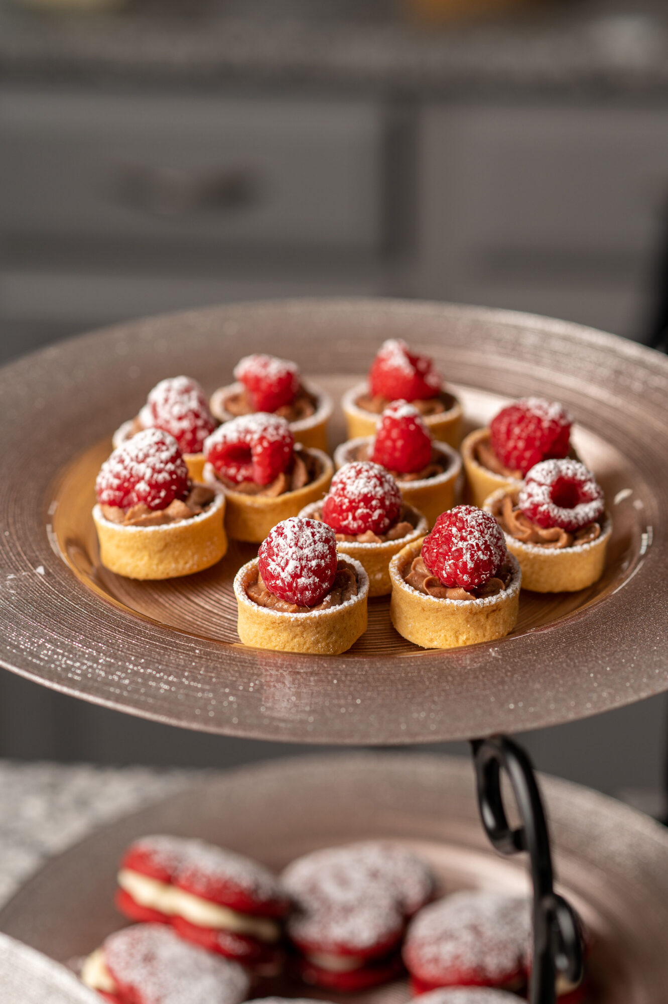 Plate with small tartlets topped with raspberries and powdered sugar, set on a table with a blurred background.