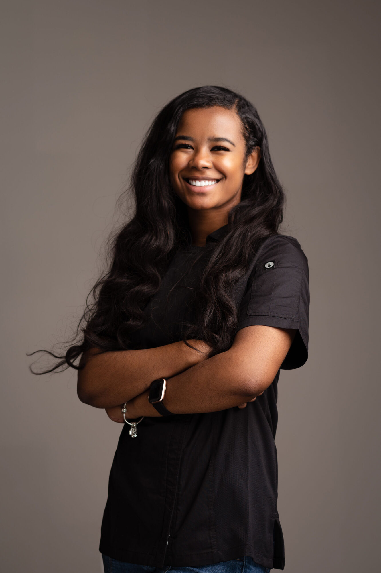 Smiling woman with long dark hair, arms crossed, wearing a black shirt, against a plain background.