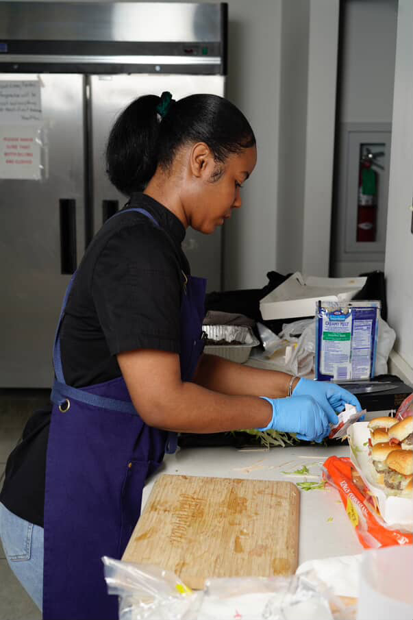 Woman wearing a black shirt and blue apron prepares food at a counter, wearing blue gloves, with kitchen appliances in background.