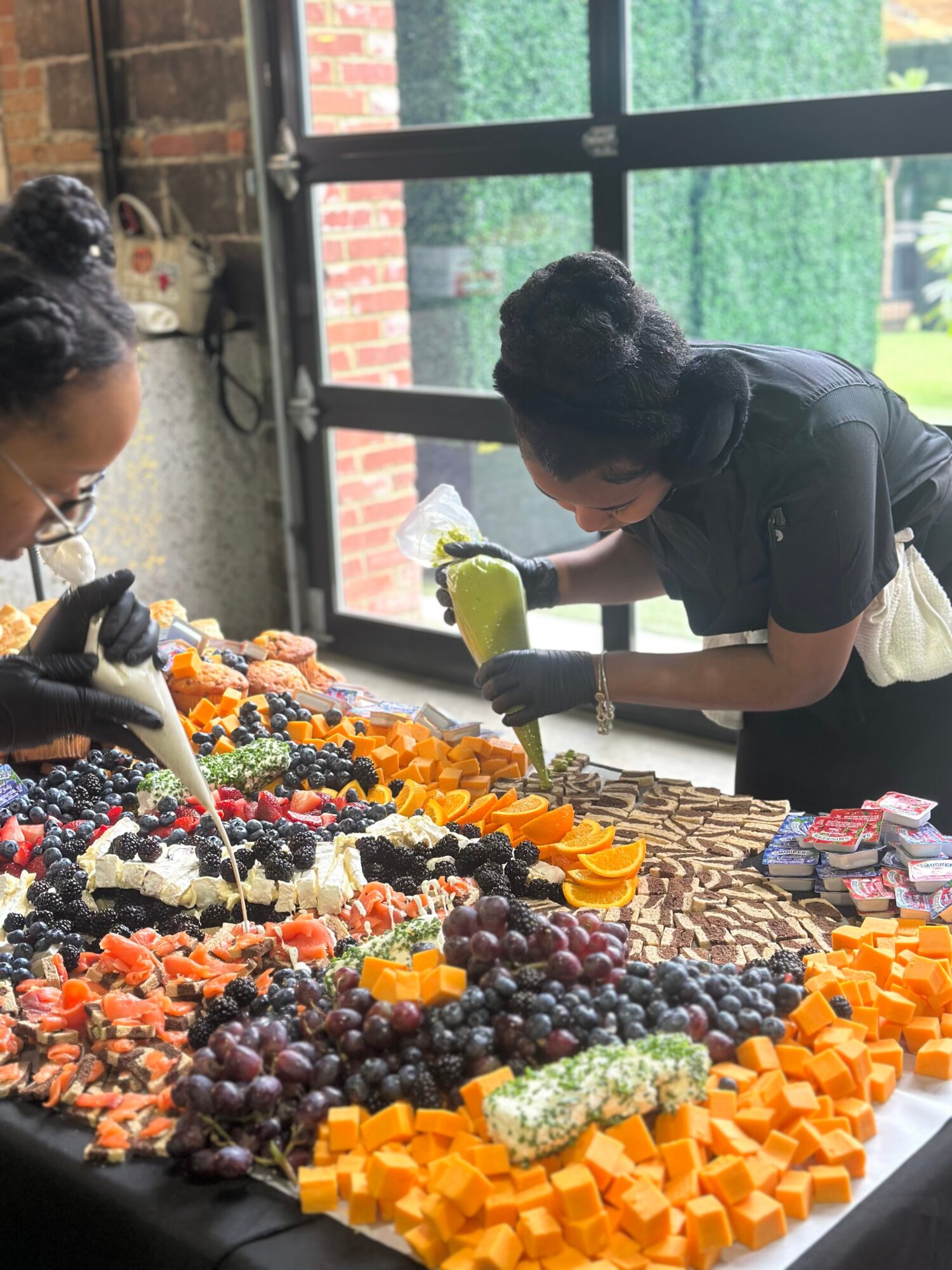 Two people decorating a large fruit and candy display on a table near a window, with various fruits and candies arranged.