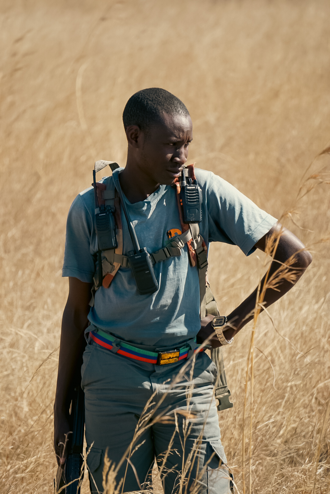 Young person with a backpack standing in a grassy field, looking down, wearing a light blue shirt and gray pants.