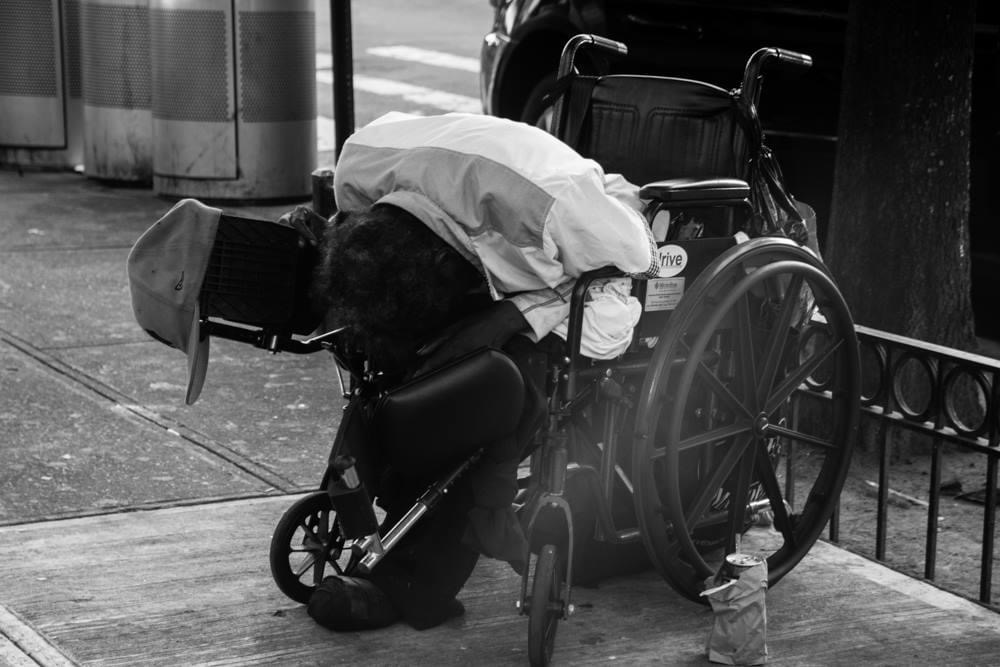Person leaning over a wheelchair on a city sidewalk, with a hat hanging on the wheelchair handle.
