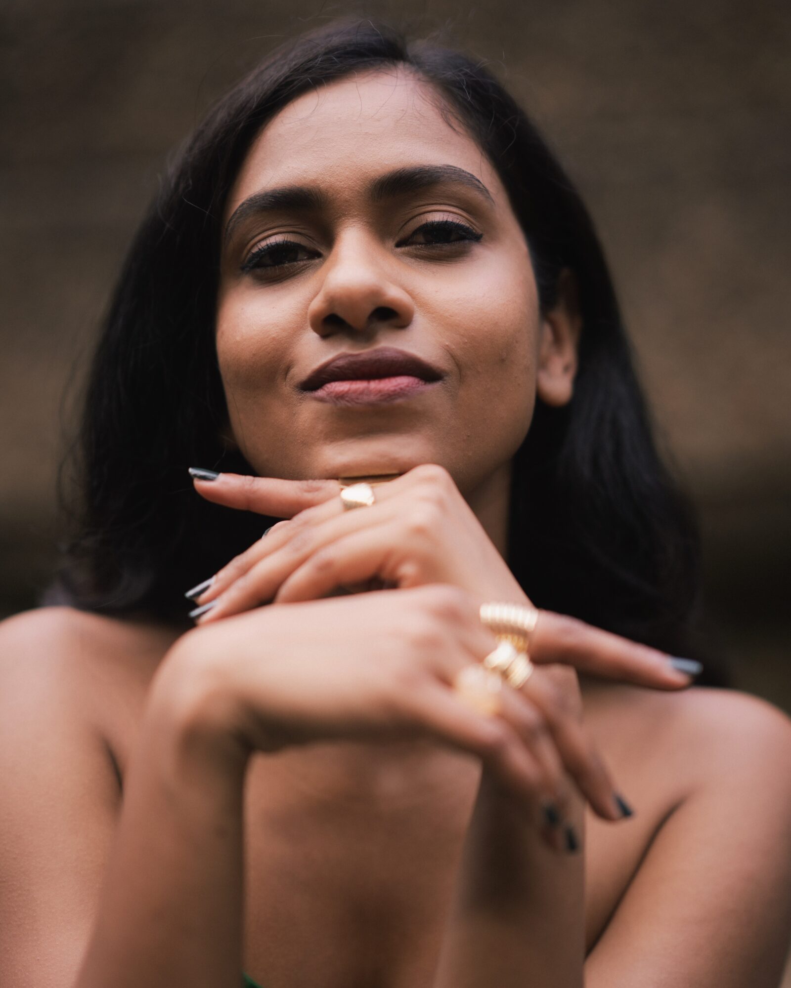 Close-up of a woman with dark hair, smiling slightly, resting her chin on her hands, wearing rings, against a blurred background.