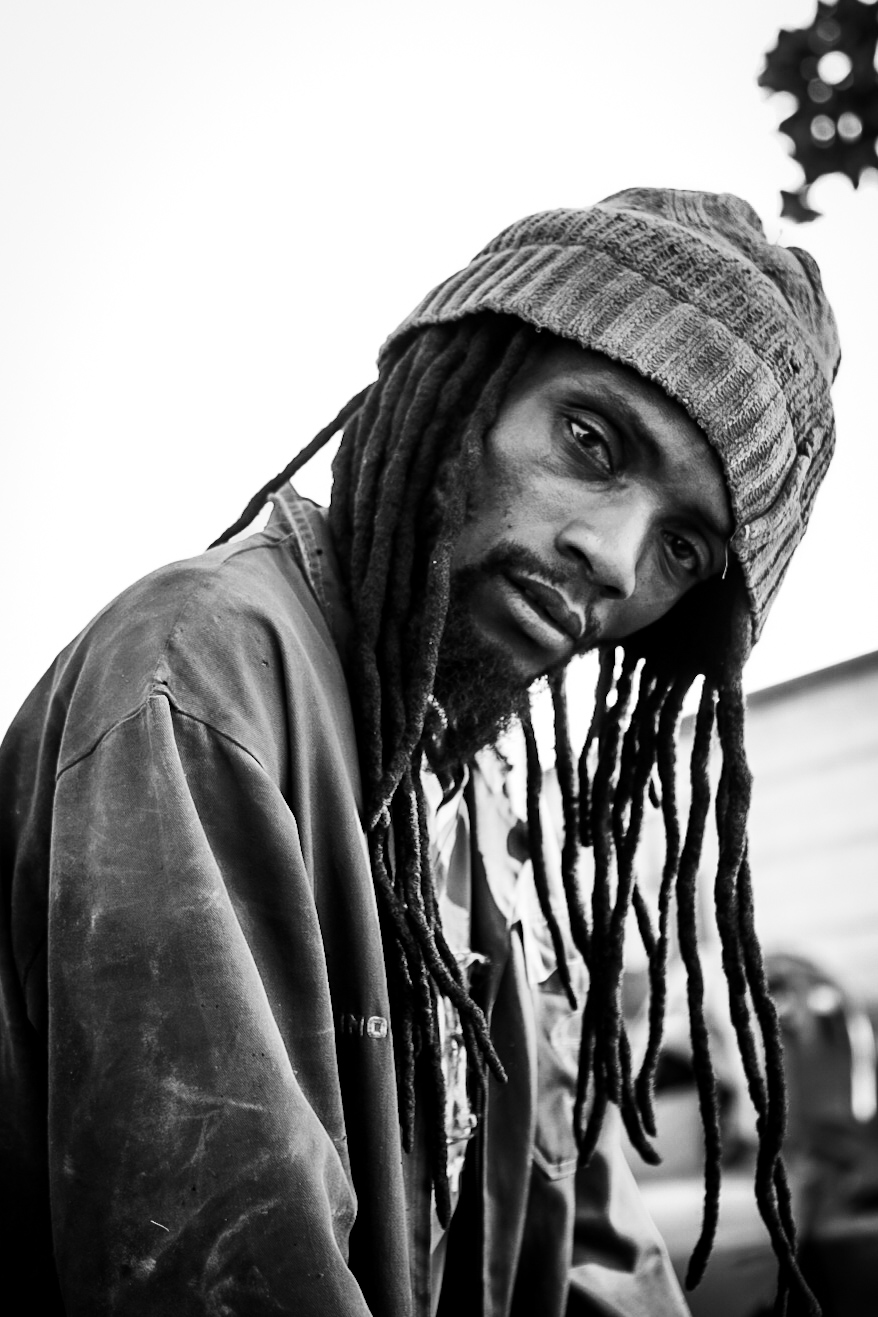 Man with dreadlocks wearing a beanie and jacket, looking at the camera, with a gear in the background.