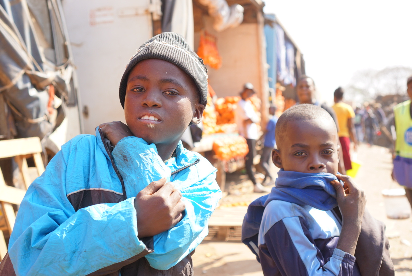 Two children stand outdoors in a busy marketplace, one wearing a gray beanie and blue jacket, the other in a dark jacket, both holding their jackets close. Word count: 20
