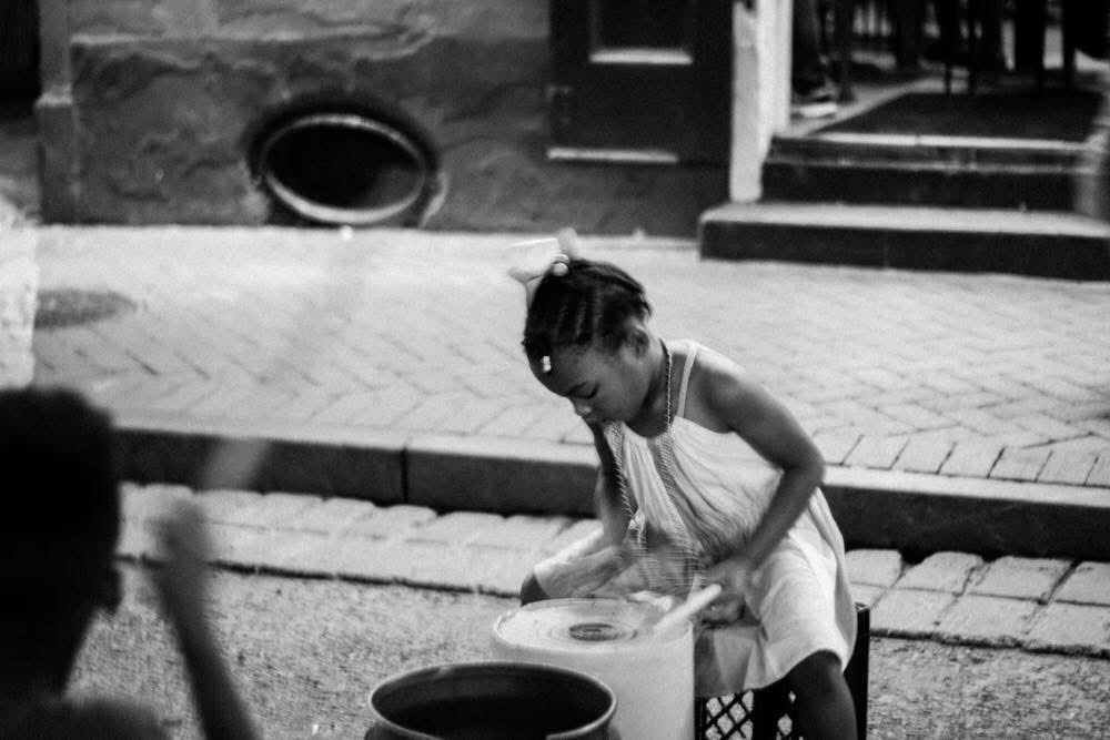 Girl in a dress crouching down, holding a plate, outdoors near steps and a building, black and white photo.