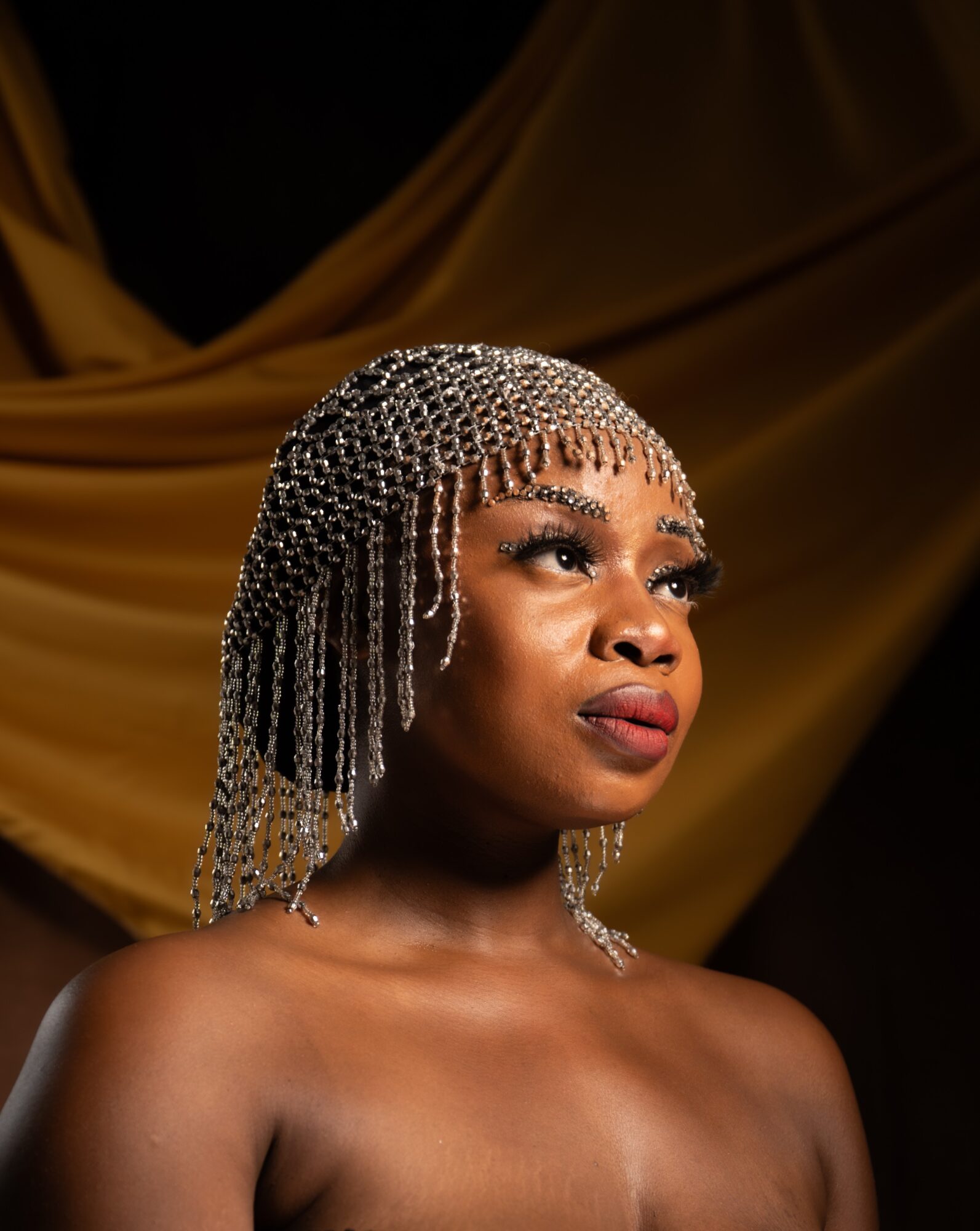 Portrait of a woman with elaborate beaded headpiece and makeup, against a gold fabric backdrop.