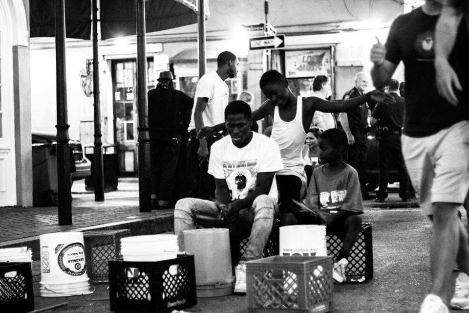 Group of people sitting and standing outdoors, some sitting on crates, with storefronts in the background, in black and white.