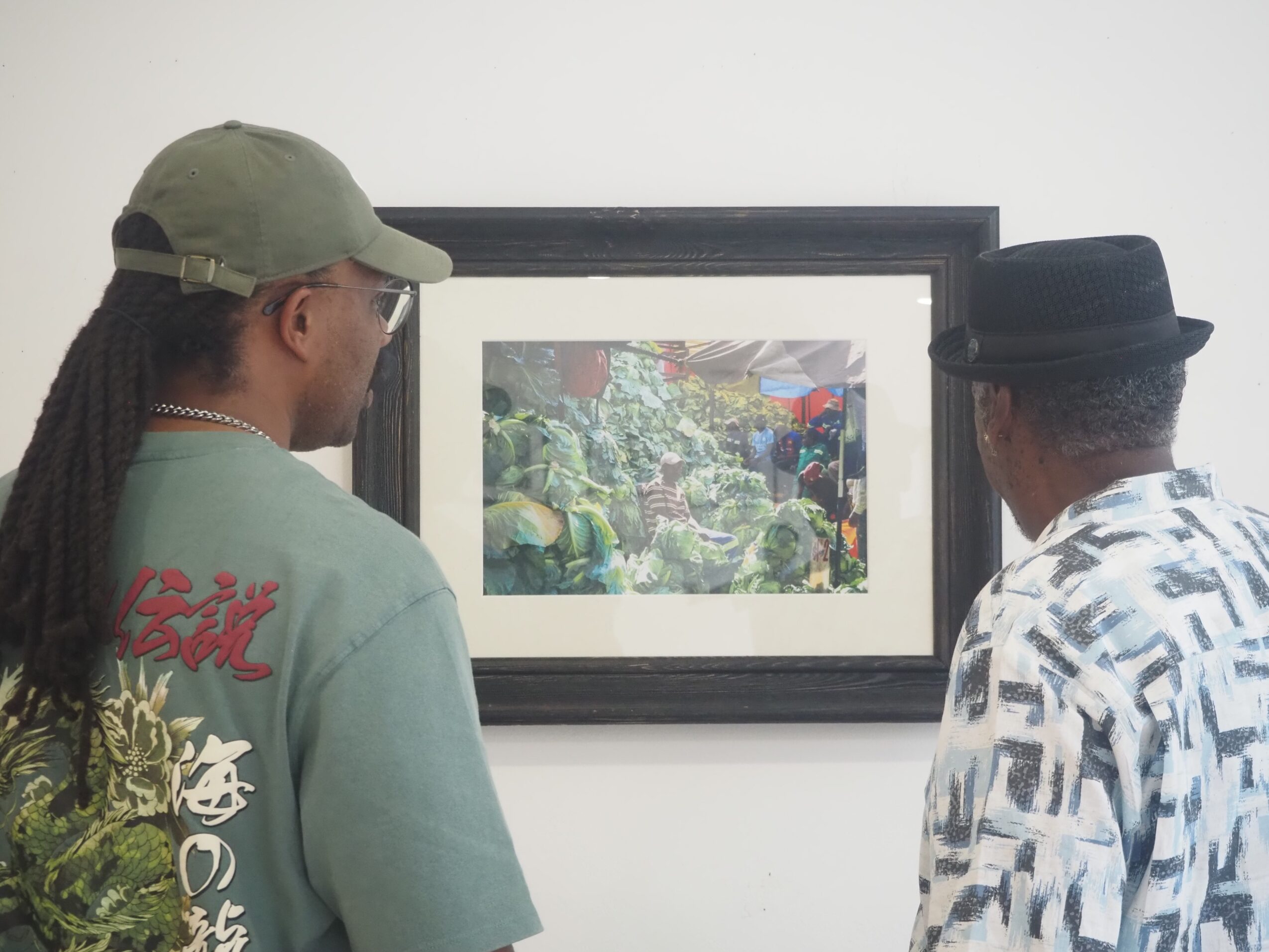 Two people viewing a framed colorful painting on a white wall, one wearing a green cap and the other a black hat.