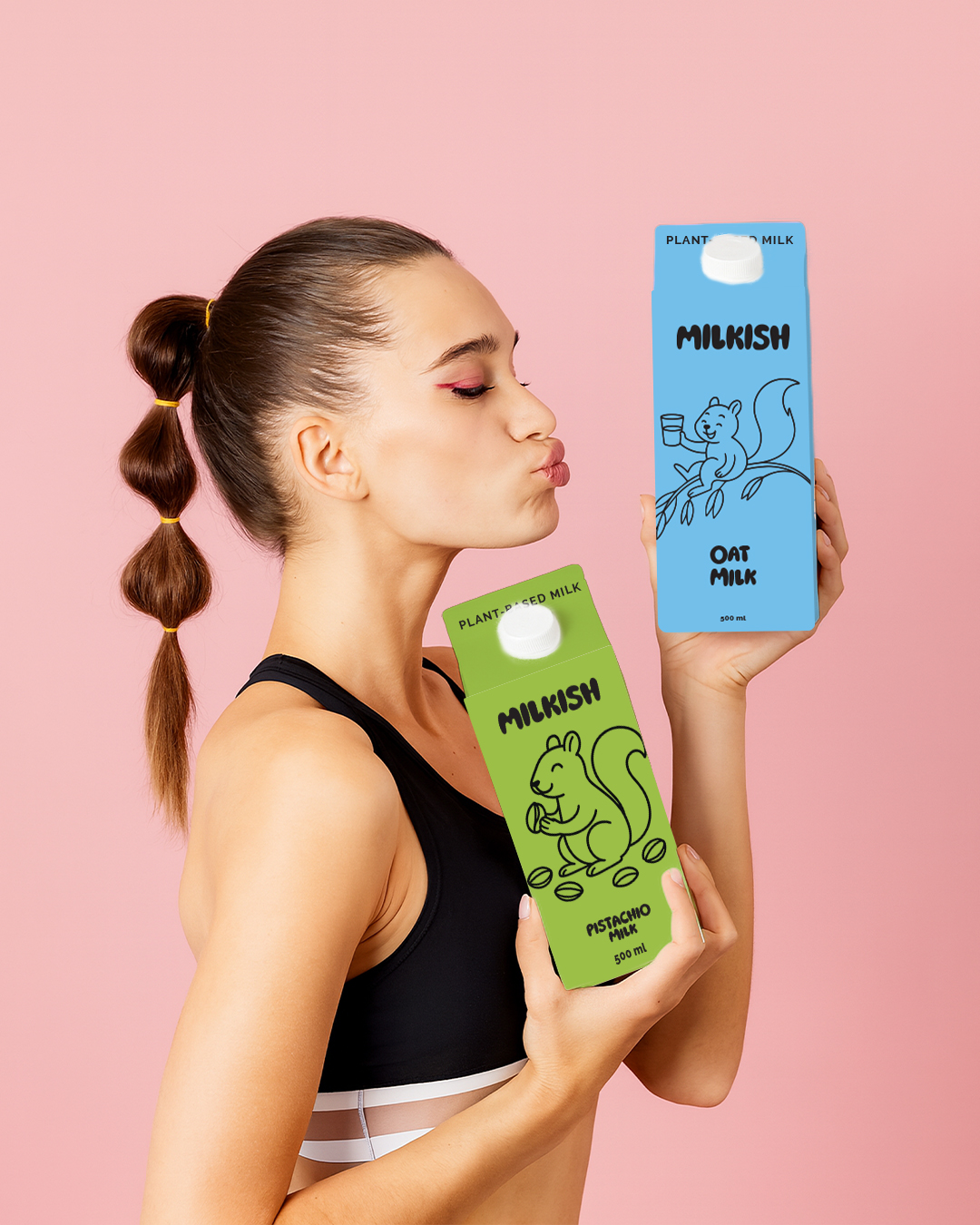 Young woman with a ponytail holding two milk cartons, one green and one blue, against a pink background.