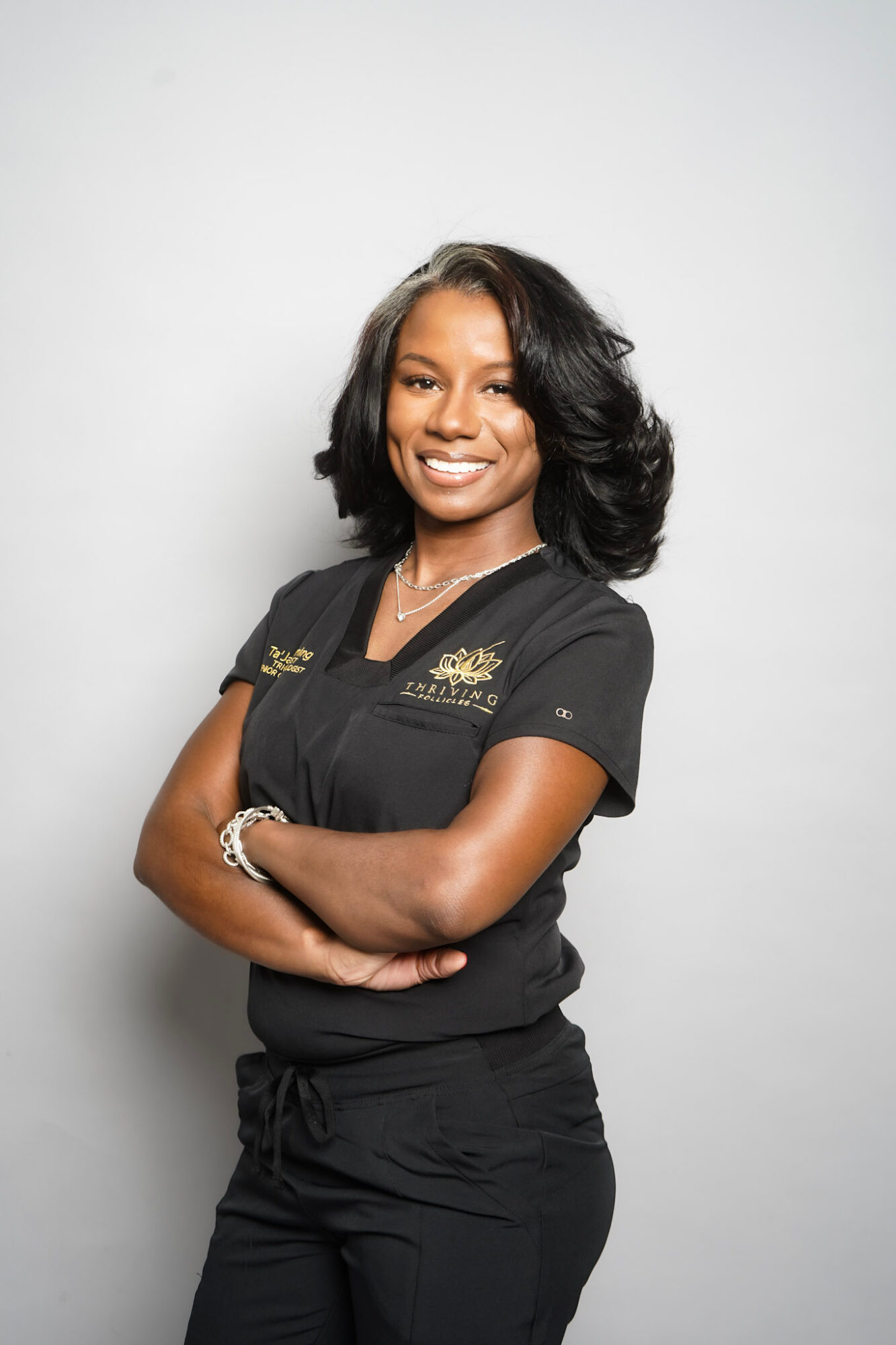 Smiling woman with shoulder-length wavy hair, wearing a black uniform with gold embroidery, arms crossed, standing against a plain white background.