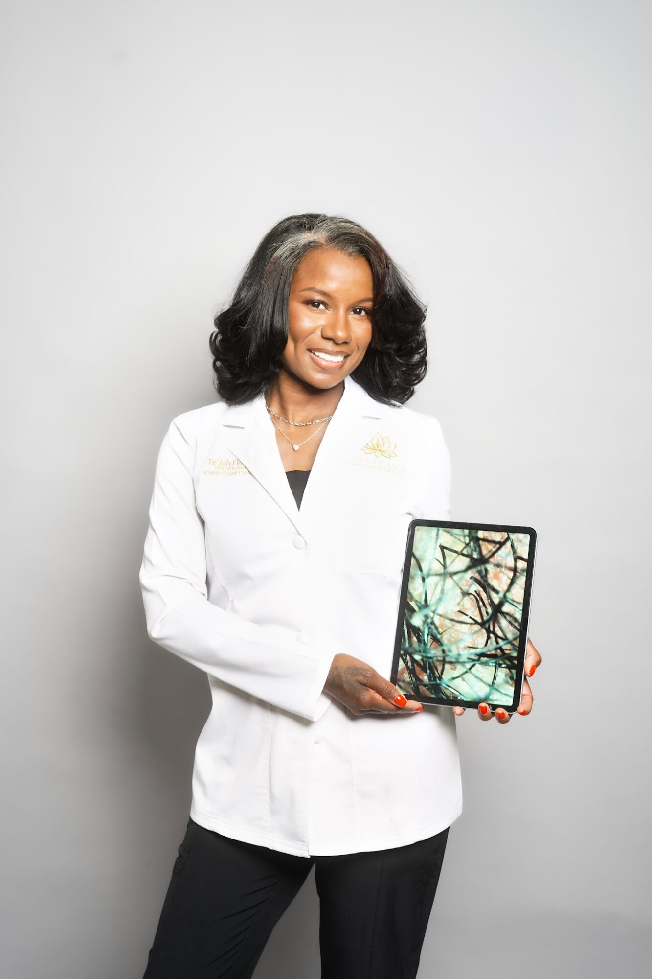 Woman in white blazer holding a tablet with a tree branch image, smiling, against a plain light background.