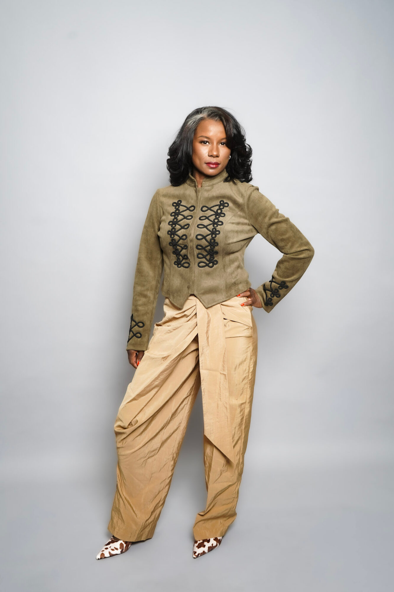 Woman with shoulder-length black hair wearing a green jacket with black embroidery and beige wide-leg pants, standing with one hand on her hip.