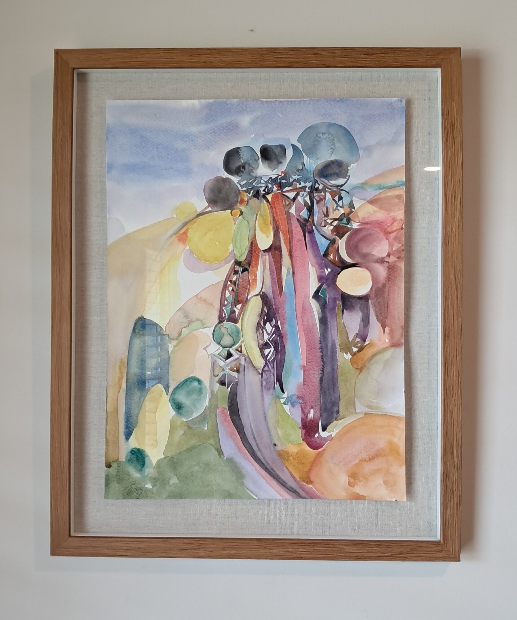 Abstract watercolor painting with colorful shapes and figures, framed in wood, hanging on a wall.