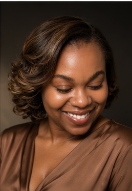 Smiling woman with styled hair and brown satin top, looking downward, dark background.