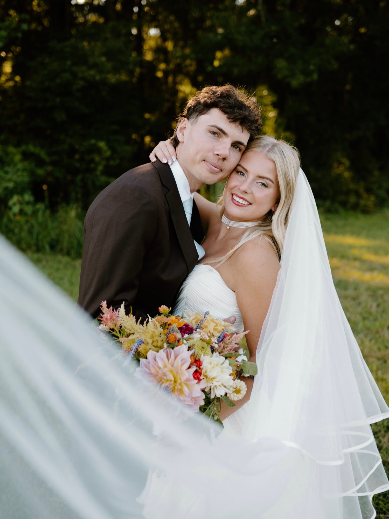 A bride and groom embrace outdoors, smiling, with trees in the background, the bride holding a colorful bouquet.