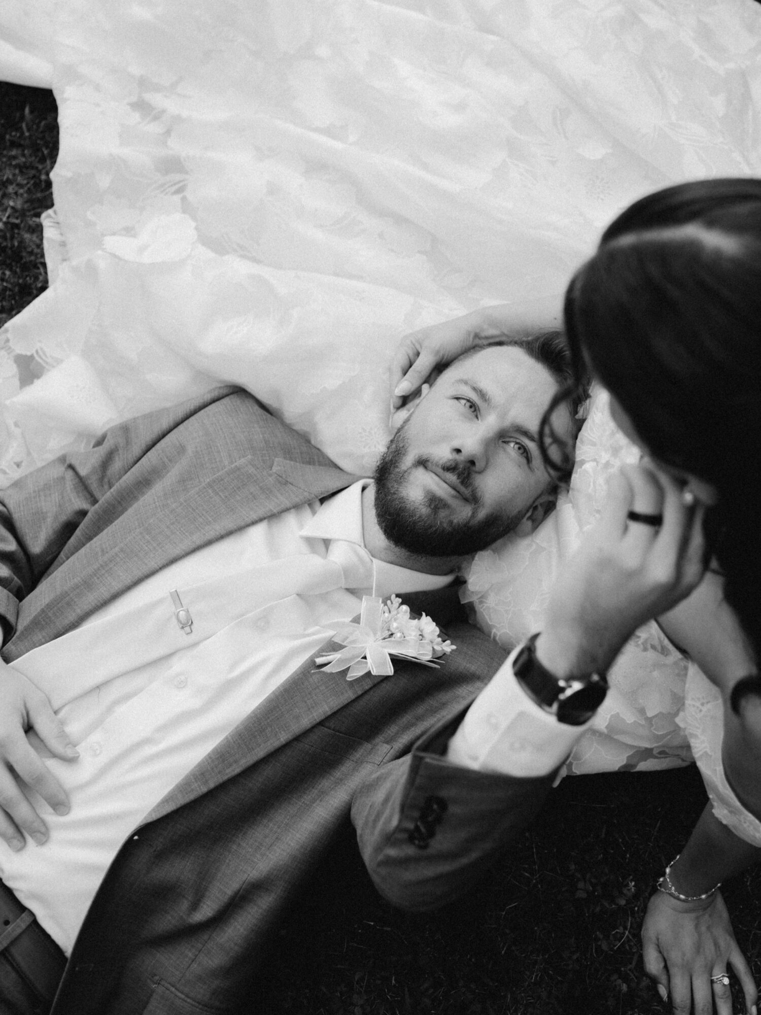 Person lying on the ground with head resting on hand, looking at another person who is leaning over them, in black and white.