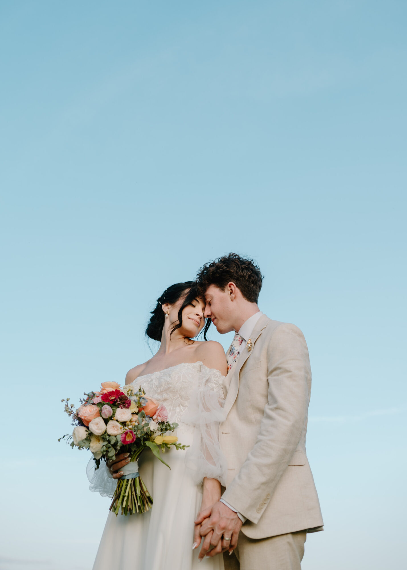 A bride and groom stand close together against a clear blue sky, holding hands, with the bride holding a bouquet of flowers.