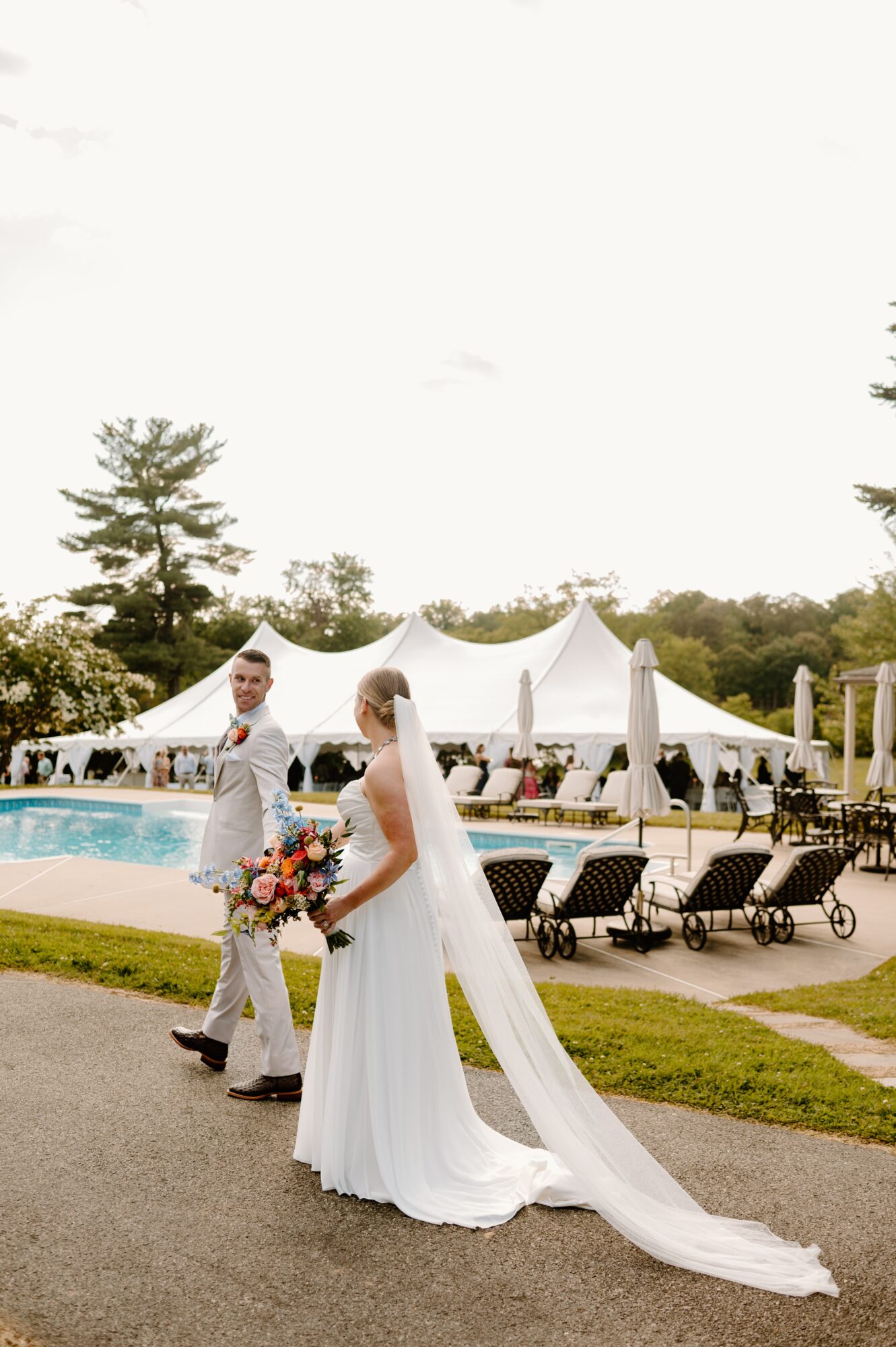 Bride and groom walking outdoors near a pool with white tents and lounge chairs, trees in the background.
