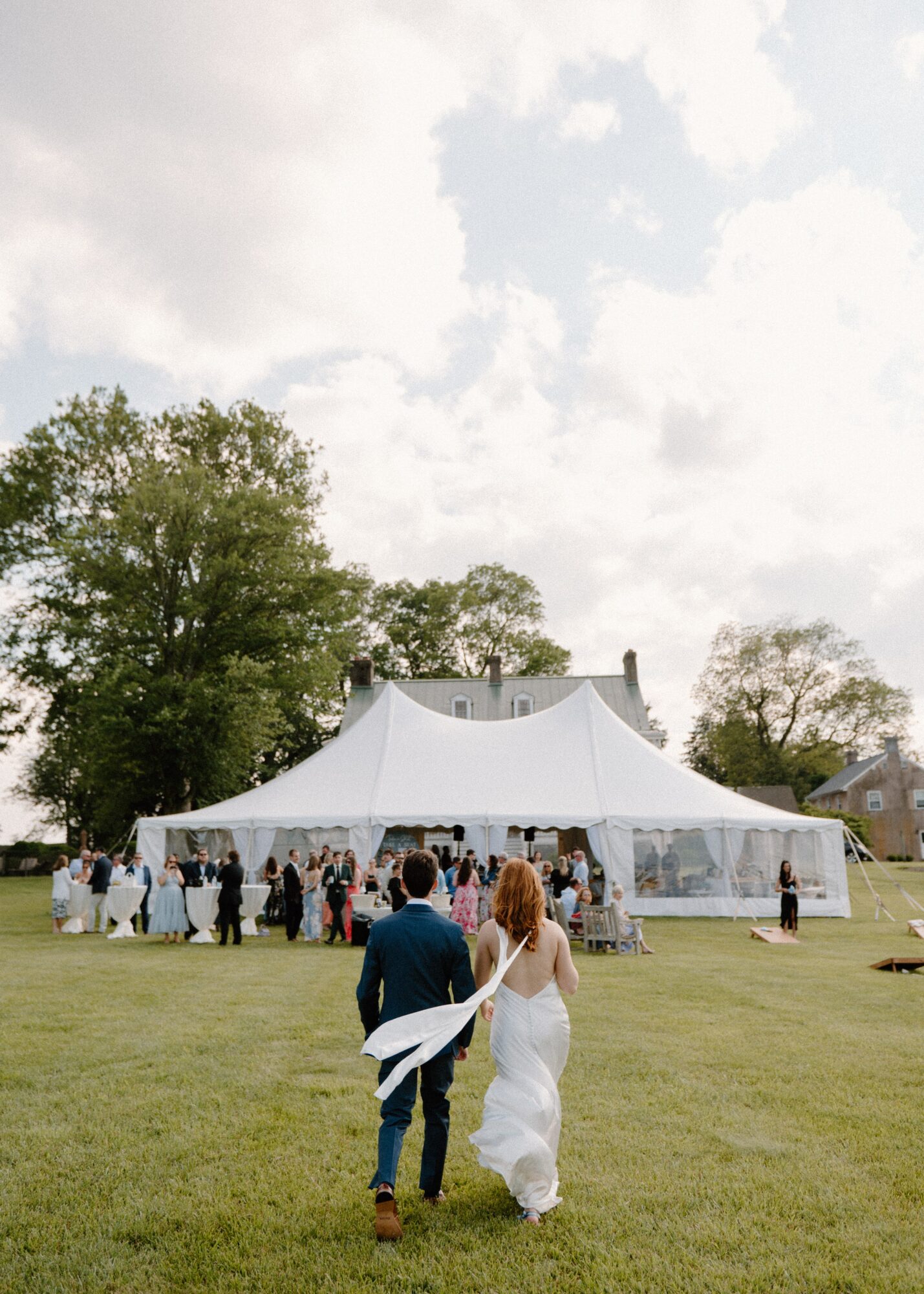 Couple walking on grass toward a large white tent during daytime outdoor event, with trees and cloudy sky in background.