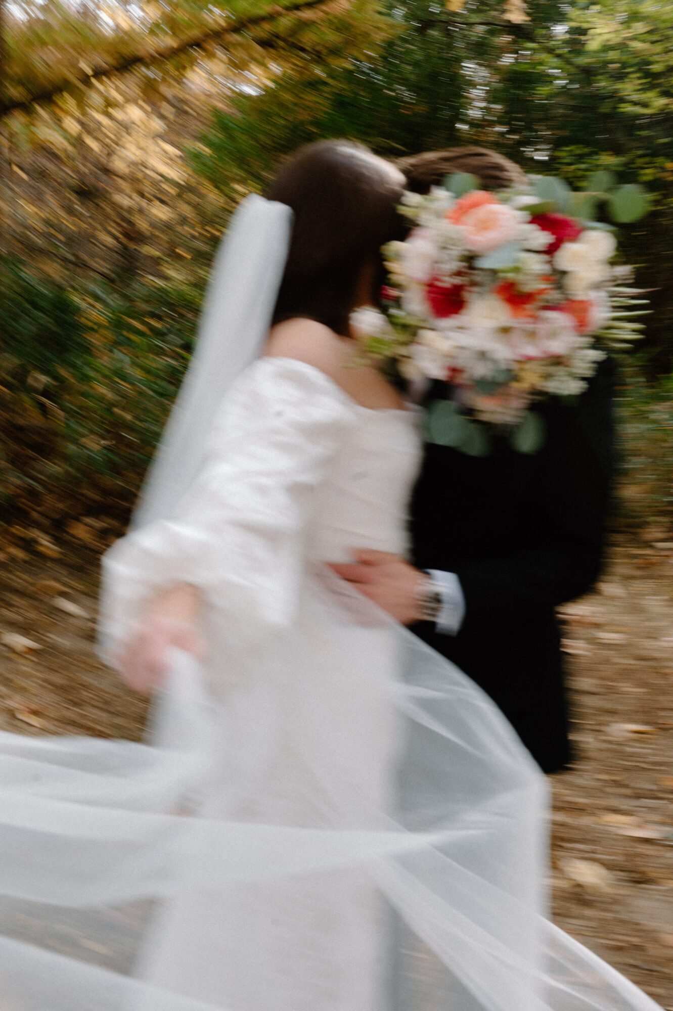 Bride and groom walking outdoors, bride holding a bouquet, surrounded by trees and fallen leaves.