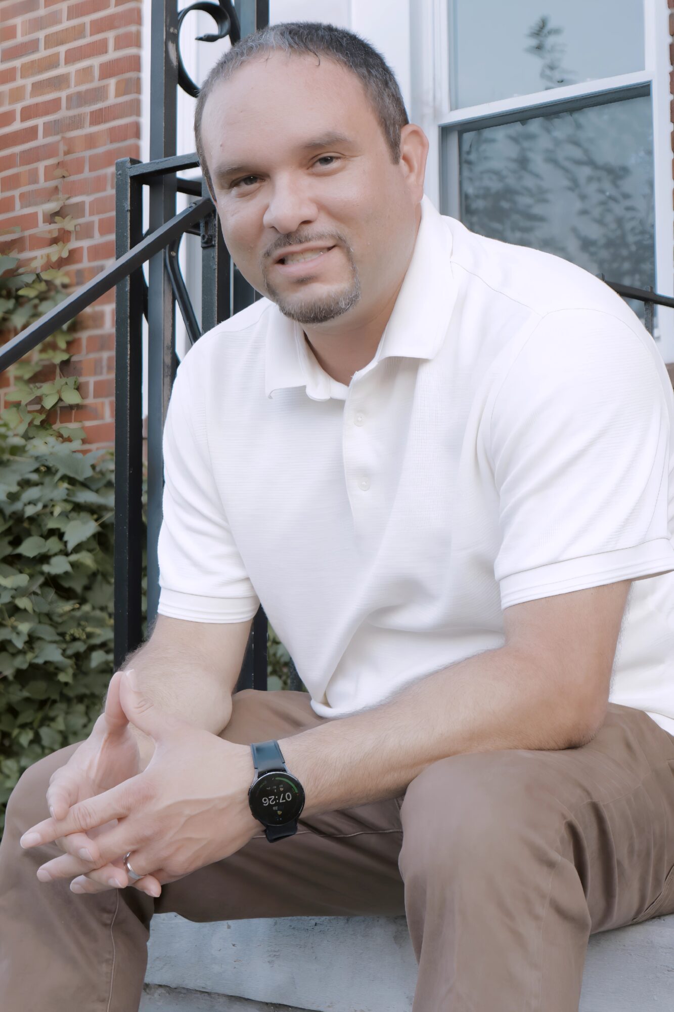 Man sitting outdoors on steps, wearing a white polo shirt and brown pants, with a brick and window background.