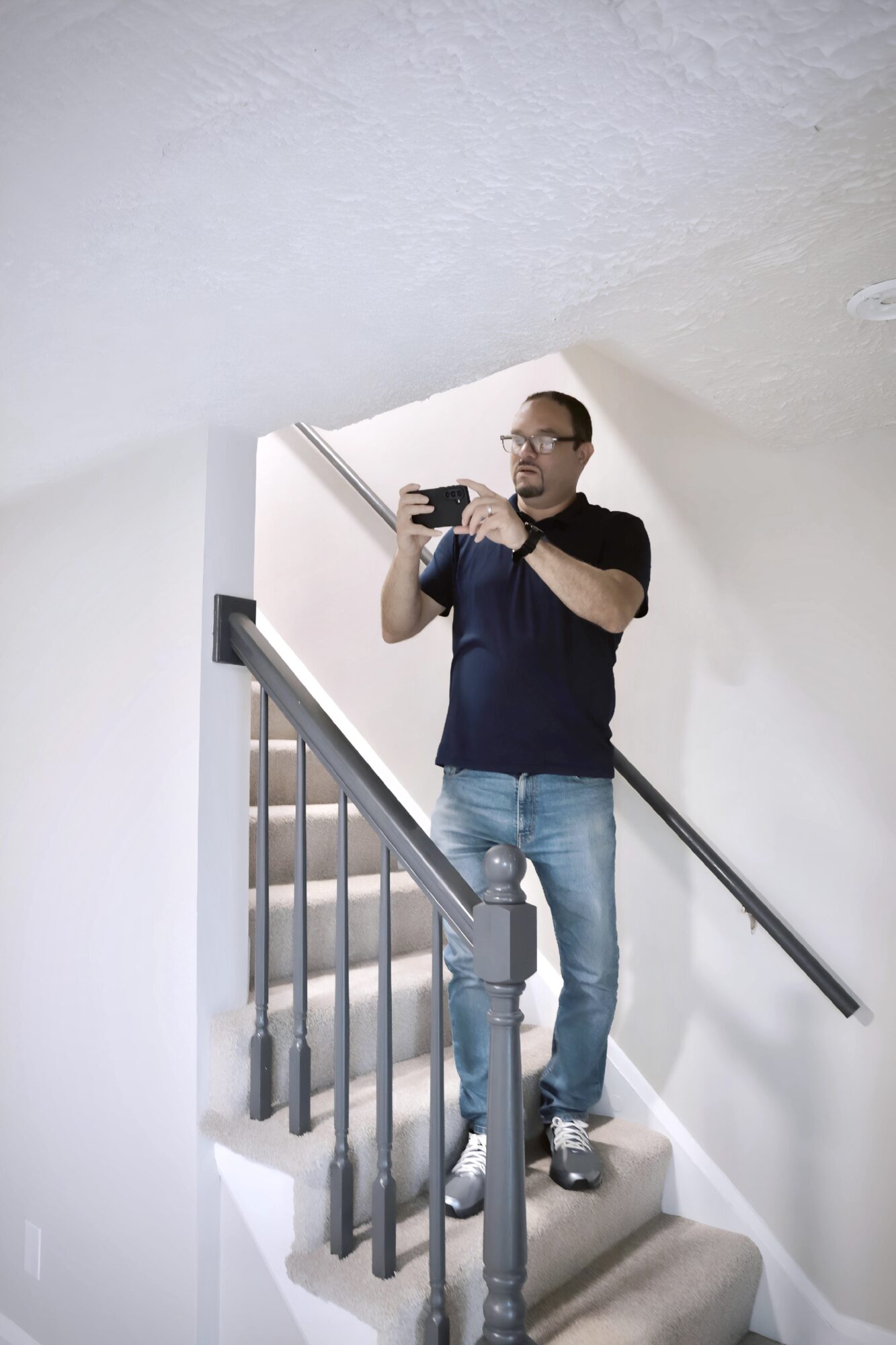 Man taking a selfie on staircase with railing, wearing glasses, black shirt, and jeans, in a white-walled room.