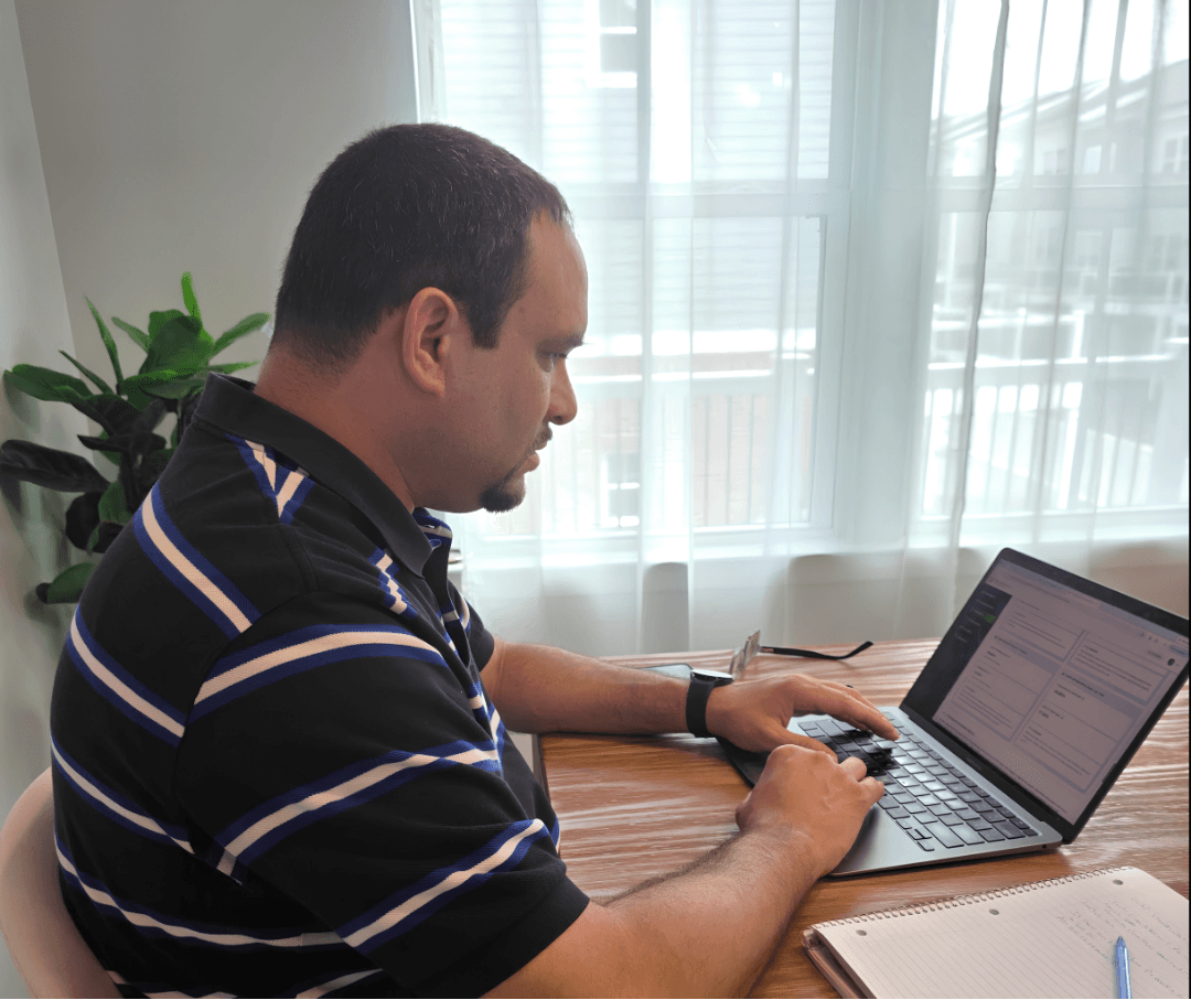 Man working on a laptop at a wooden table with a notebook, in a room with large windows and curtains