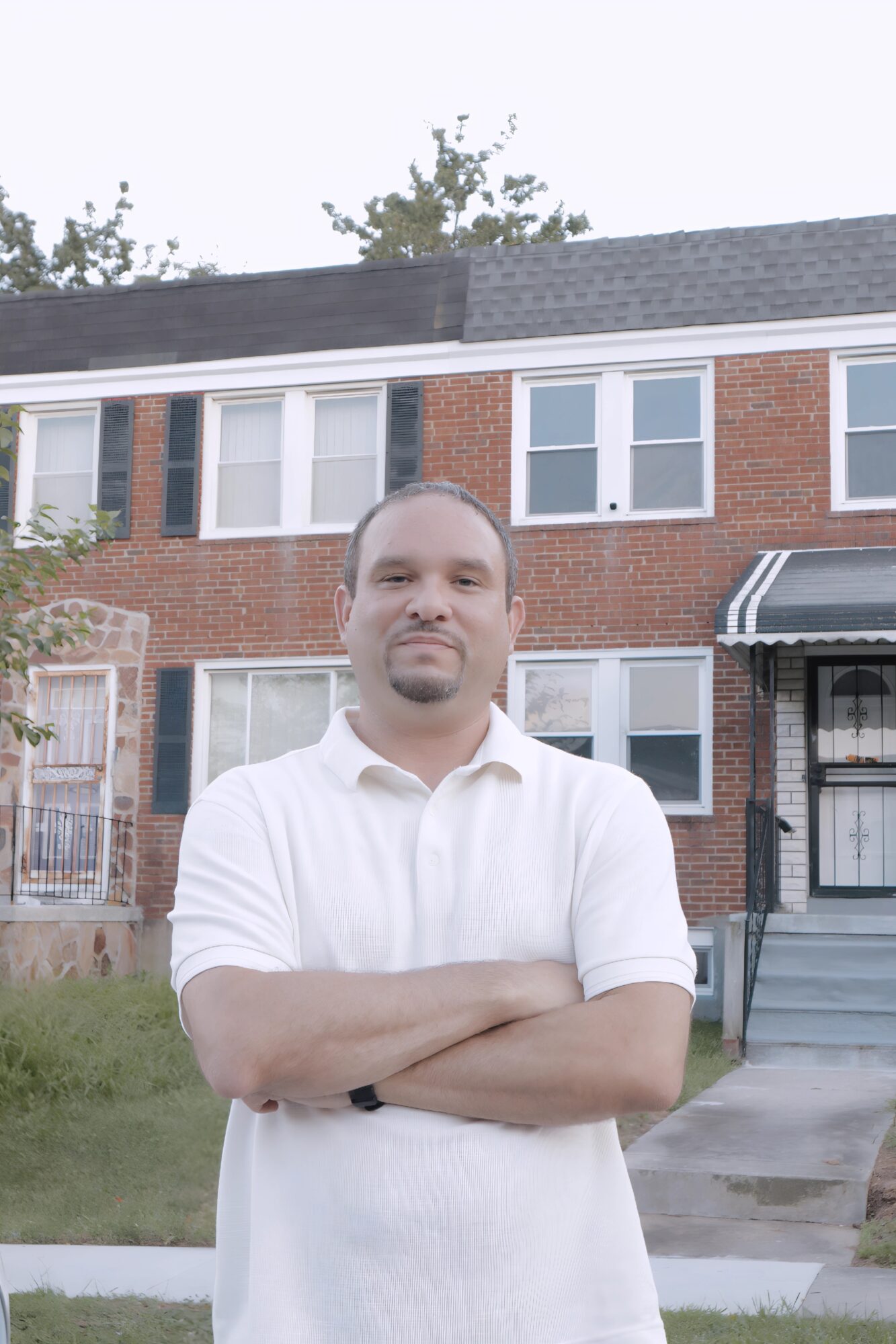 Man with crossed arms standing outside in front of a brick residential building, wearing a white shirt.