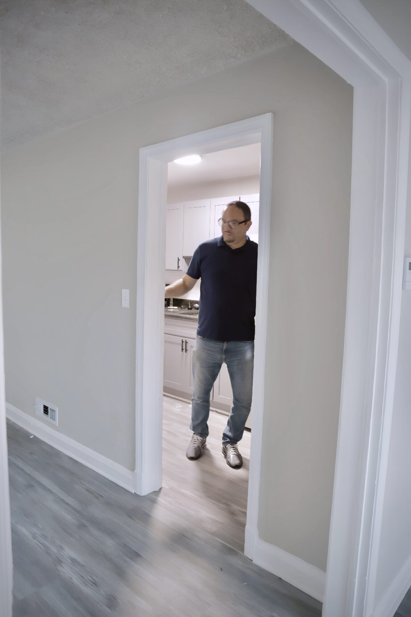 Man standing in doorway of kitchen, wearing glasses, black shirt, jeans, and sneakers, looking to the side.
