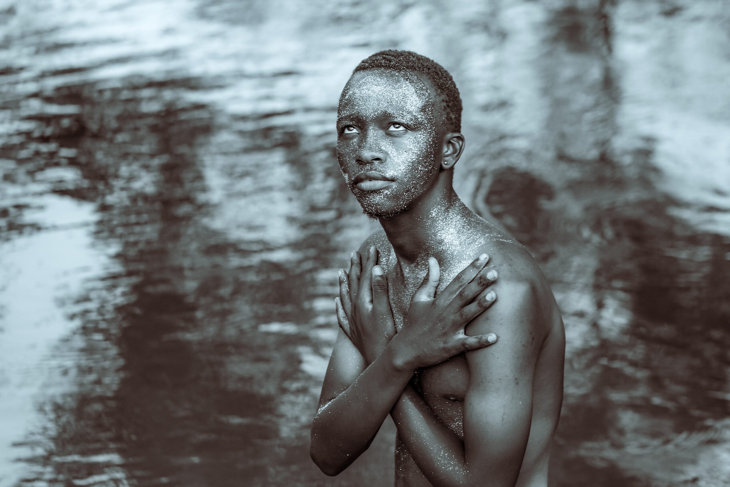 Young person with short hair and bare shoulders stands near water, crossing arms over chest, looking to the side.