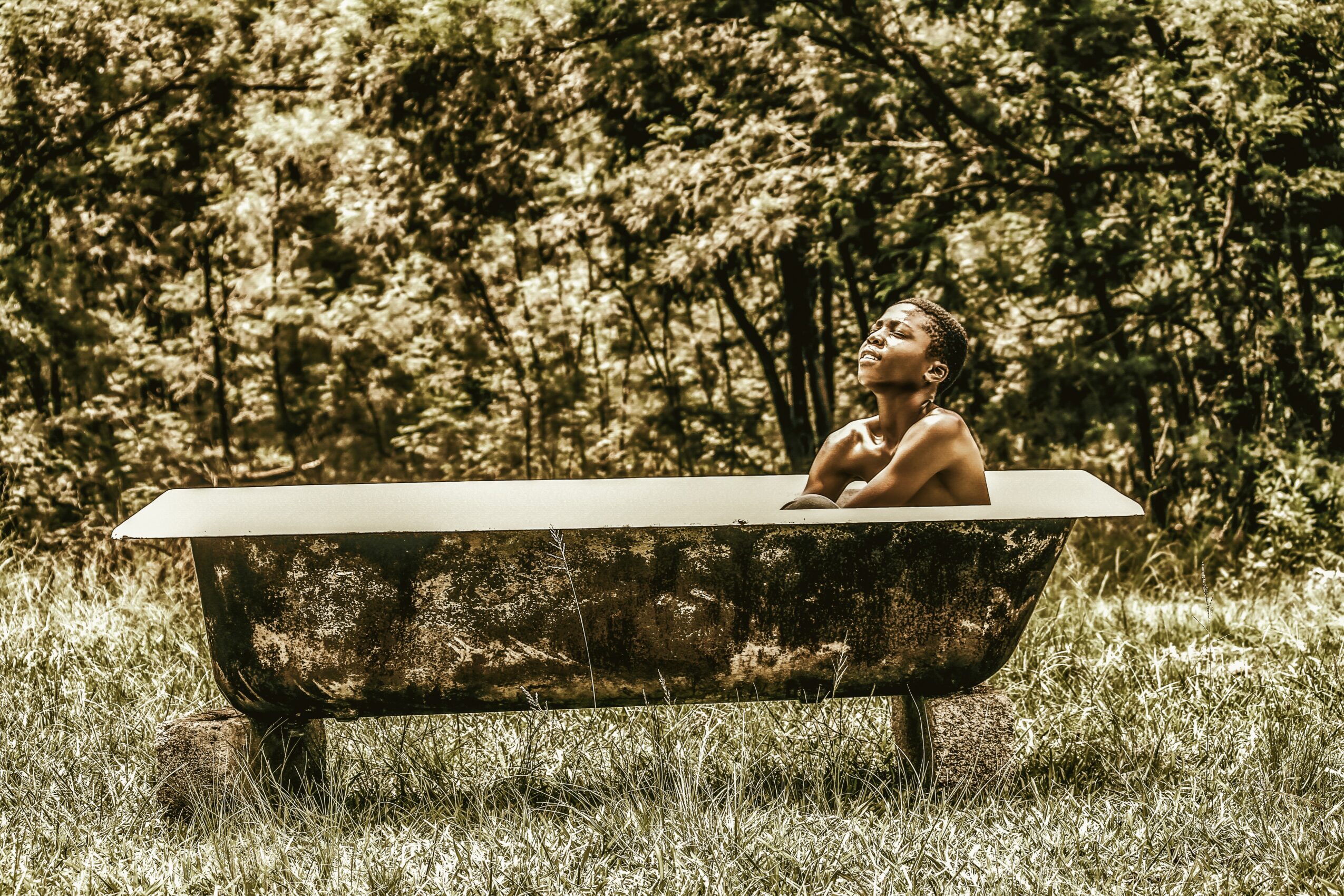 Person sitting in a bathtub outdoors in a grassy area with trees in the background, looking upward with hand near face.