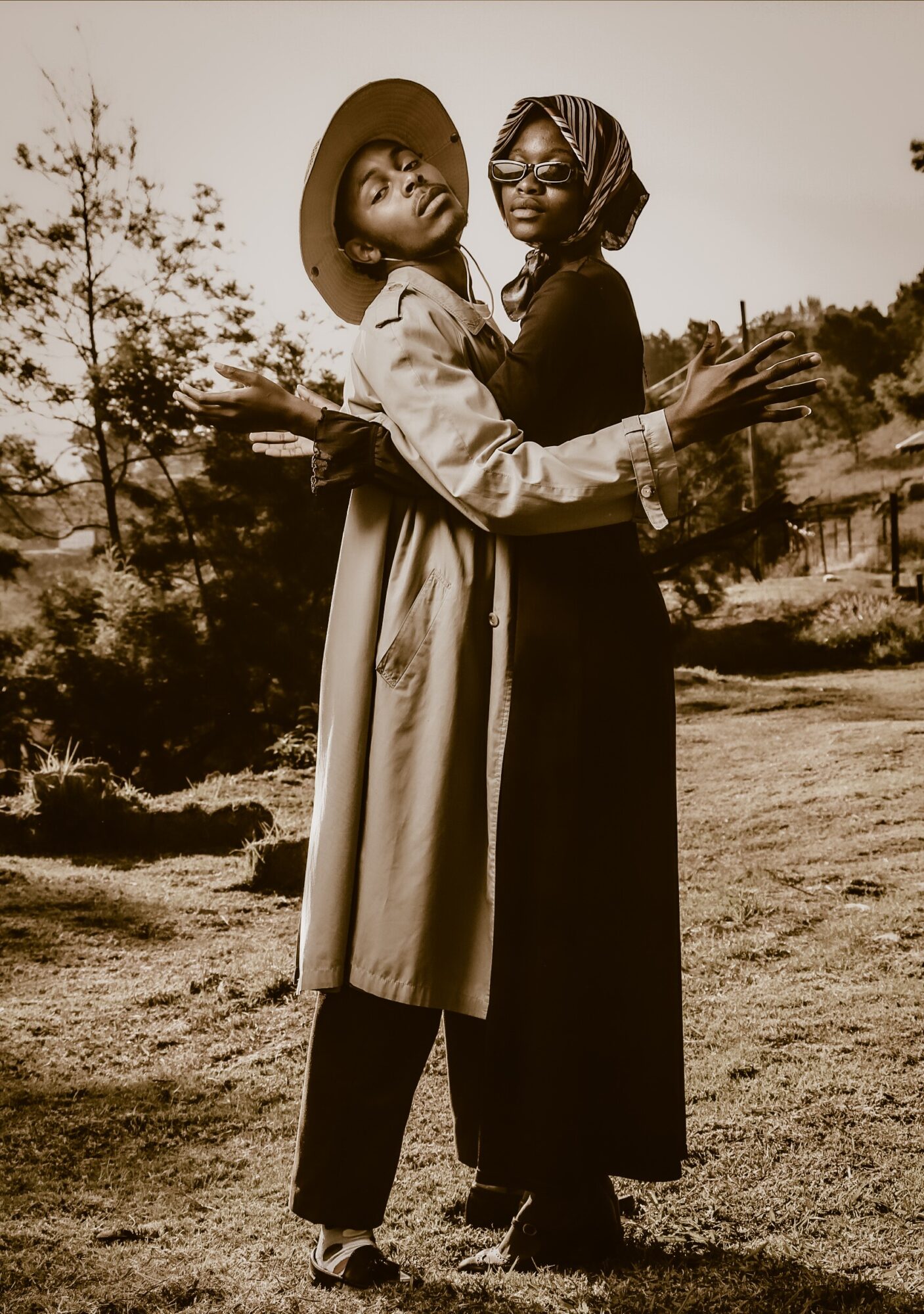 Two women embrace outdoors, one wearing a wide-brimmed hat and trench coat, the other with glasses and headscarf, in a natural setting.