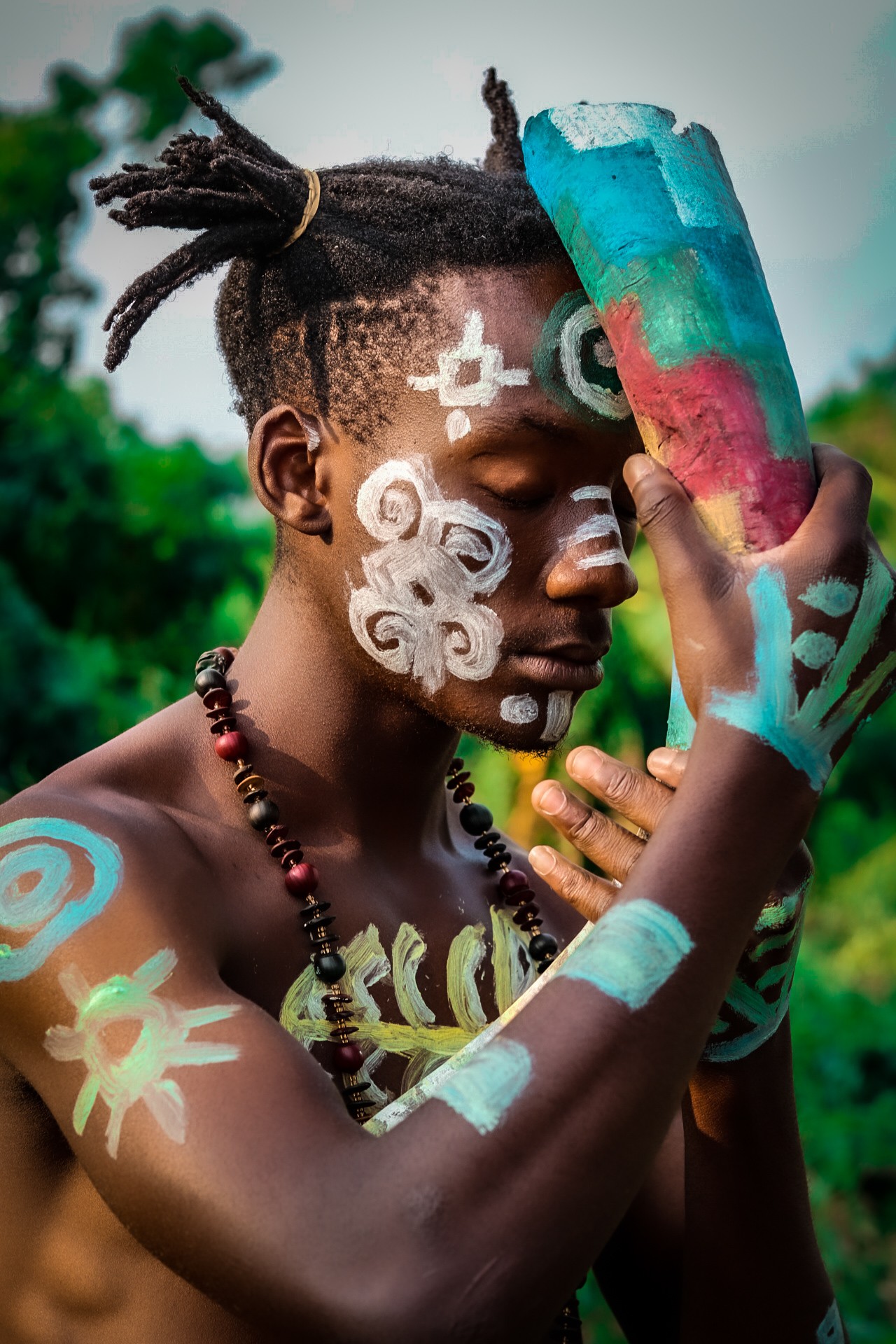 Young person with traditional face and body paint, holding a colorful object, outdoors with greenery in background.