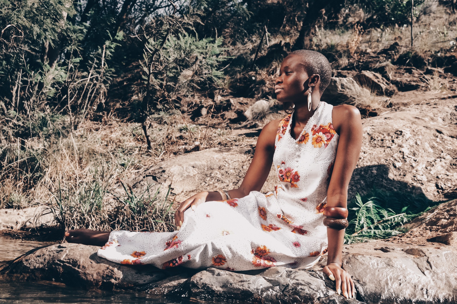 Woman sitting on a rock outdoors, wearing a white dress with floral embroidery, surrounded by natural scenery.