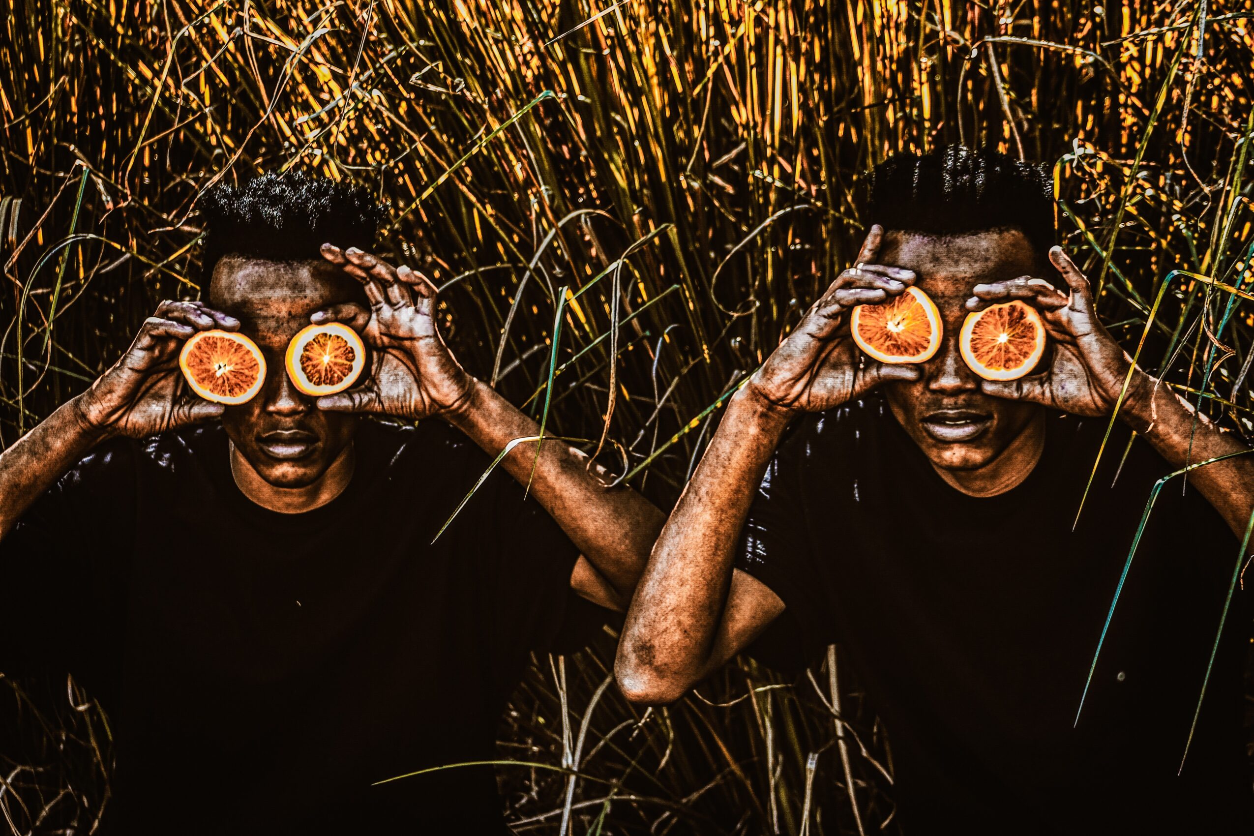 Two people hold halved oranges in front of their eyes, surrounded by tall grass or reeds, in a natural setting.