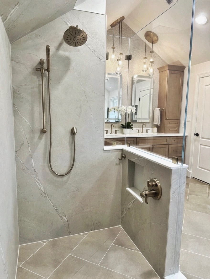 Bathroom shower with a handheld showerhead, marble wall, and a partial view of the vanity and mirror in the background.