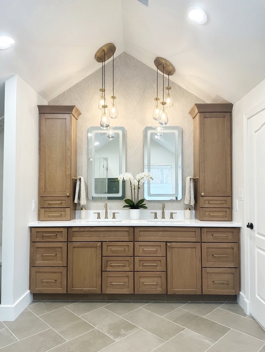 Bathroom vanity with two mirrors, wooden cabinets, and a white countertop with a flower arrangement, under a sloped ceiling with pendant lights.
