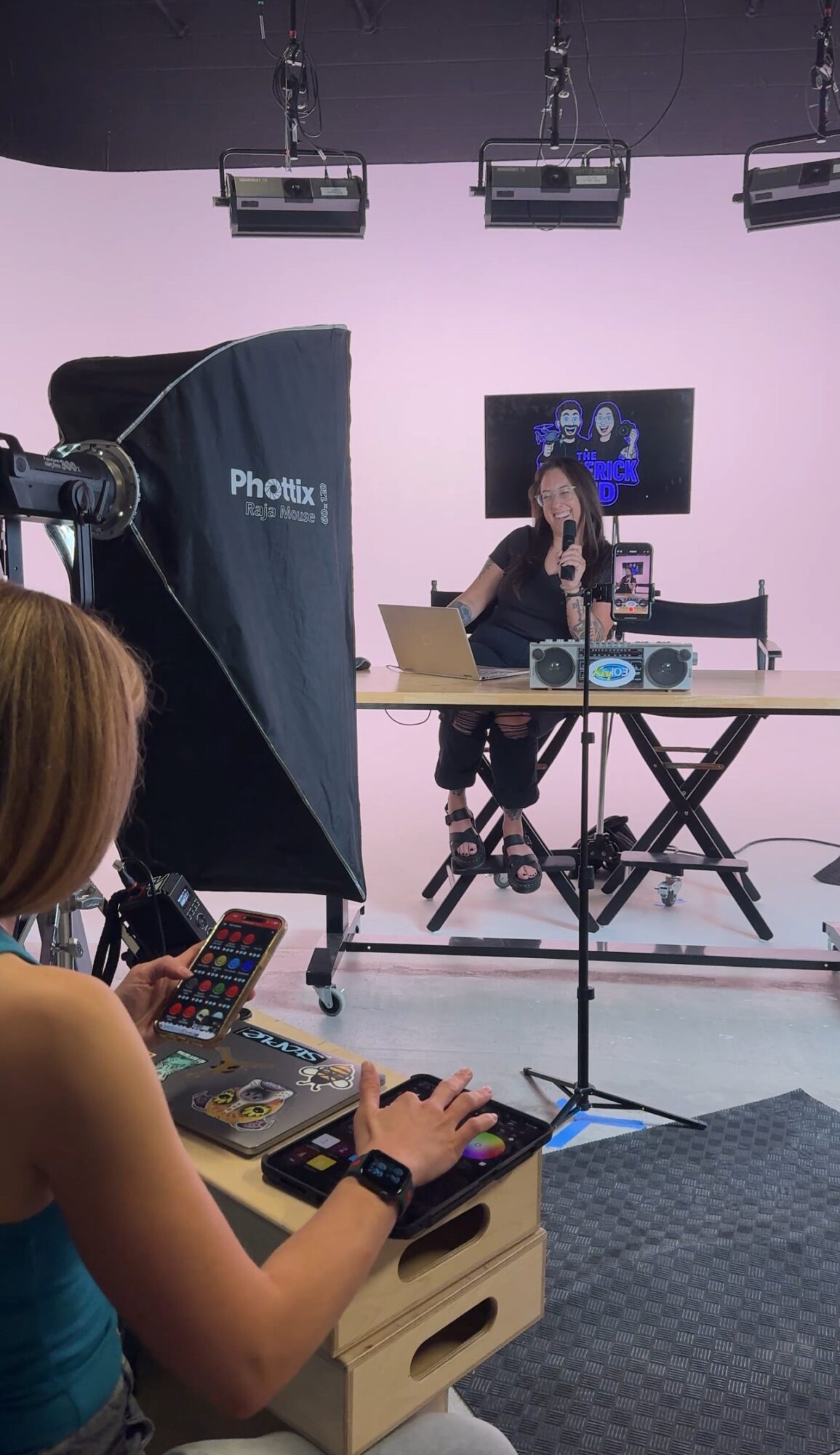 A woman with a remote controls a DJ setup in a studio with a presenter on a screen behind her.