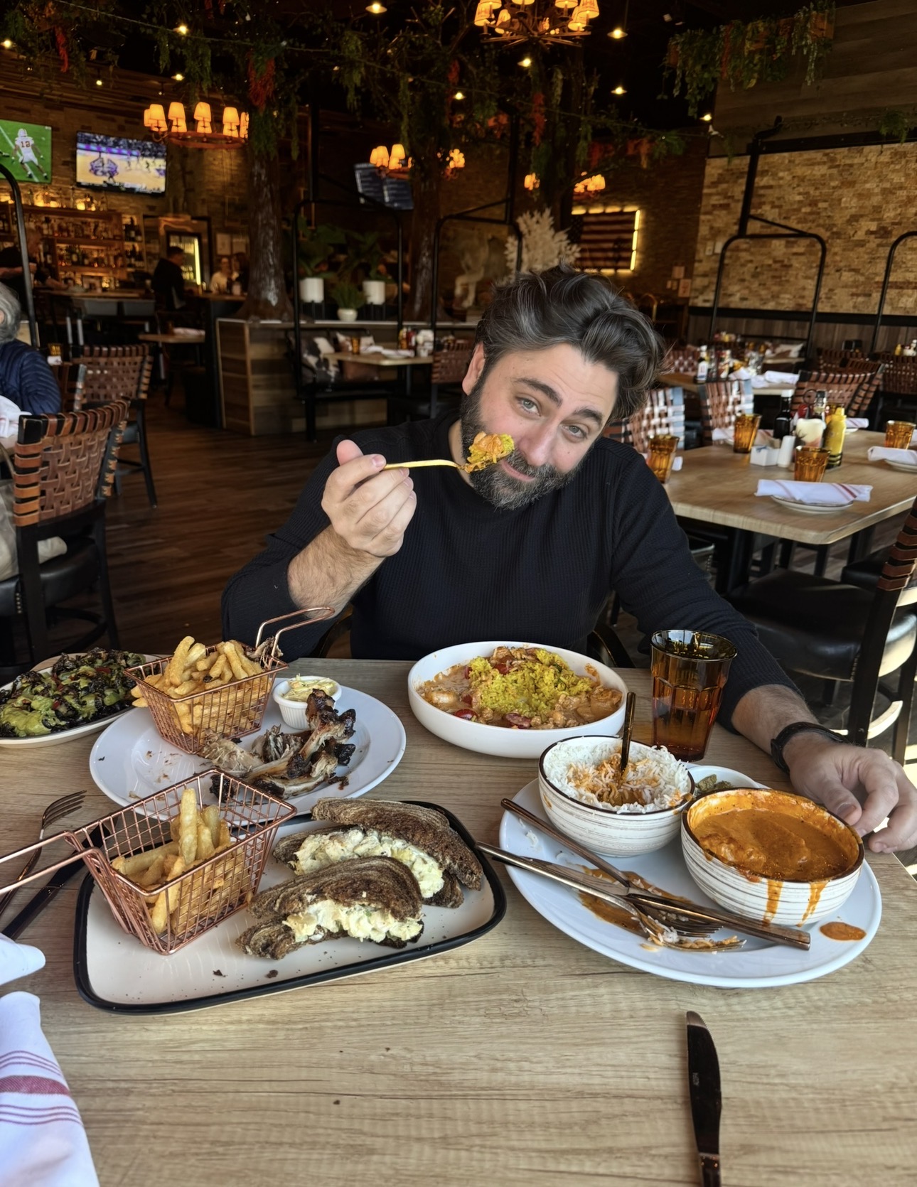 Person with dark hair and beard eating at a restaurant table with various dishes, holding a fork, smiling, in a cozy setting.