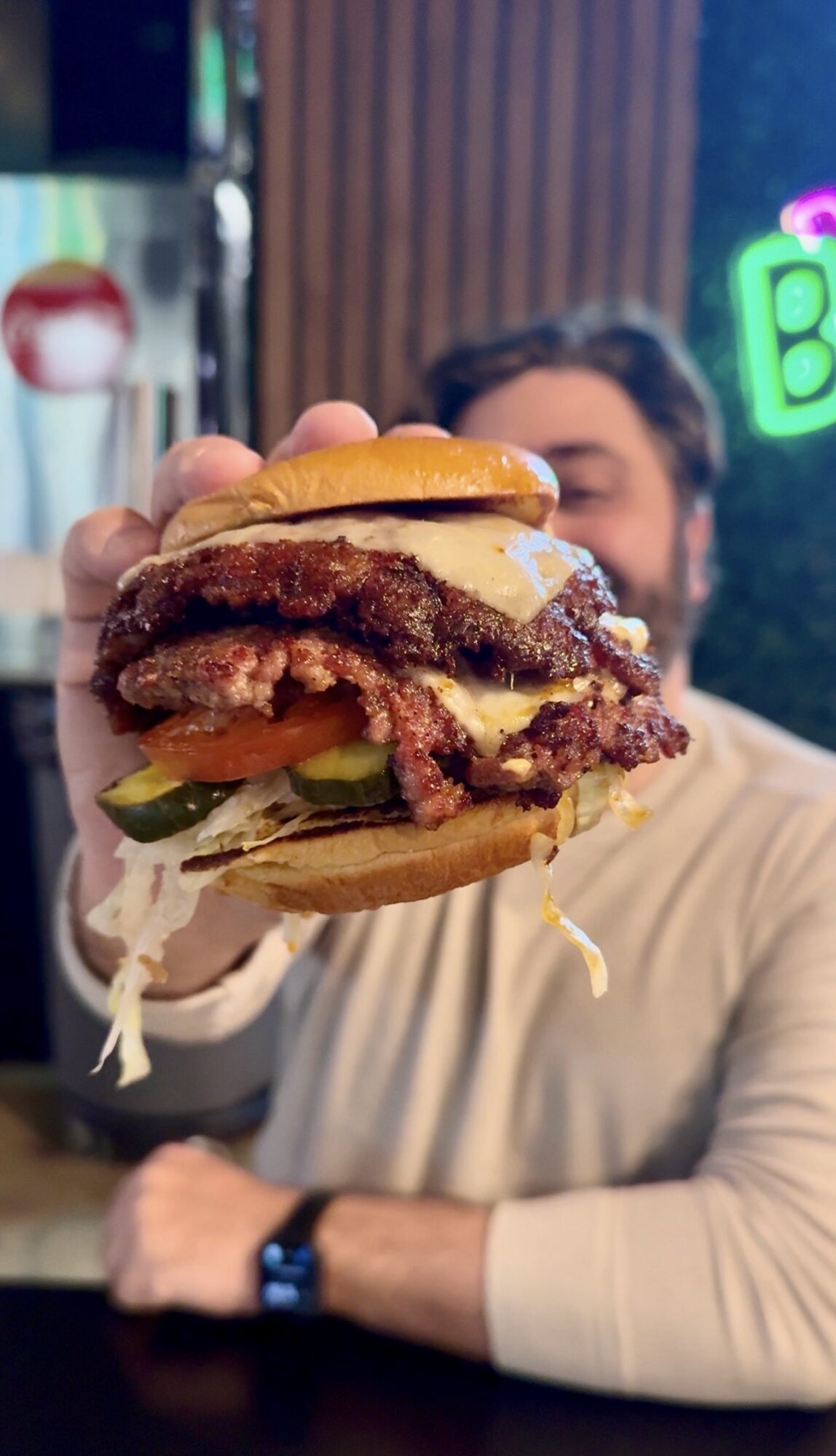 Person holding a burger with lettuce, tomato, pickles, and a beef patty, in a restaurant setting with neon lights.