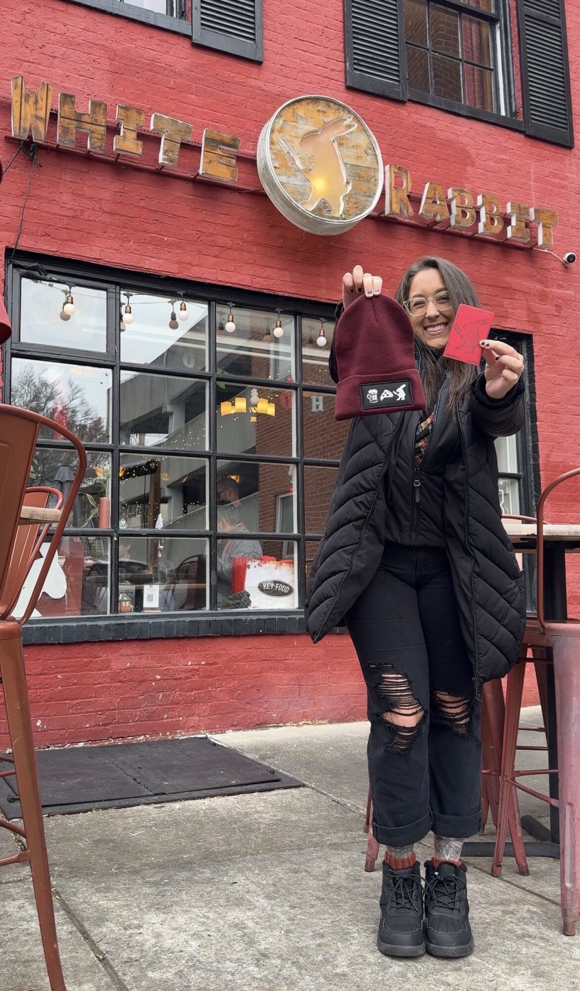 Person standing outside a red building holding a drink and a phone, smiling, with large windows reflecting the street.